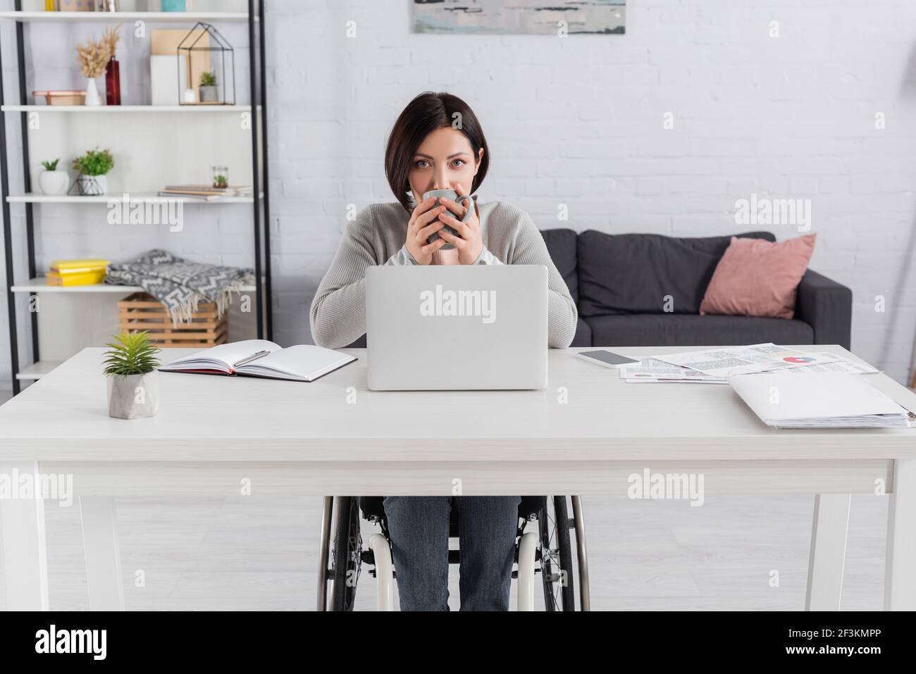 Donna handicappata che beve caffè vicino a gadget e carte sul tavolo Foto Stock