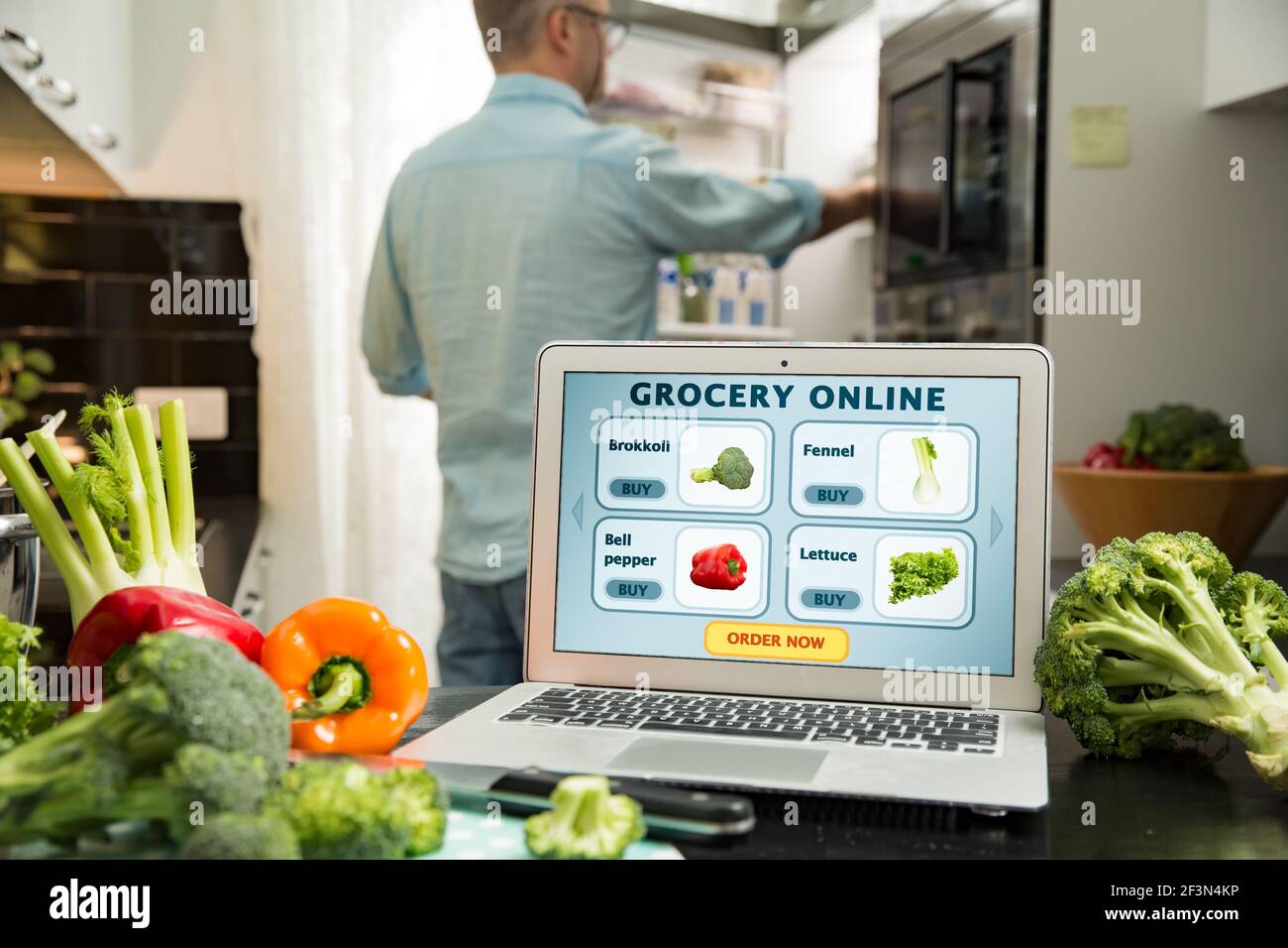 Uomo che usa il computer portatile e cucina in cucina a casa. Acquisto di generi alimentari online. Pagina web del negozio online di alimenti sullo schermo verdure fresche in cucina Foto Stock