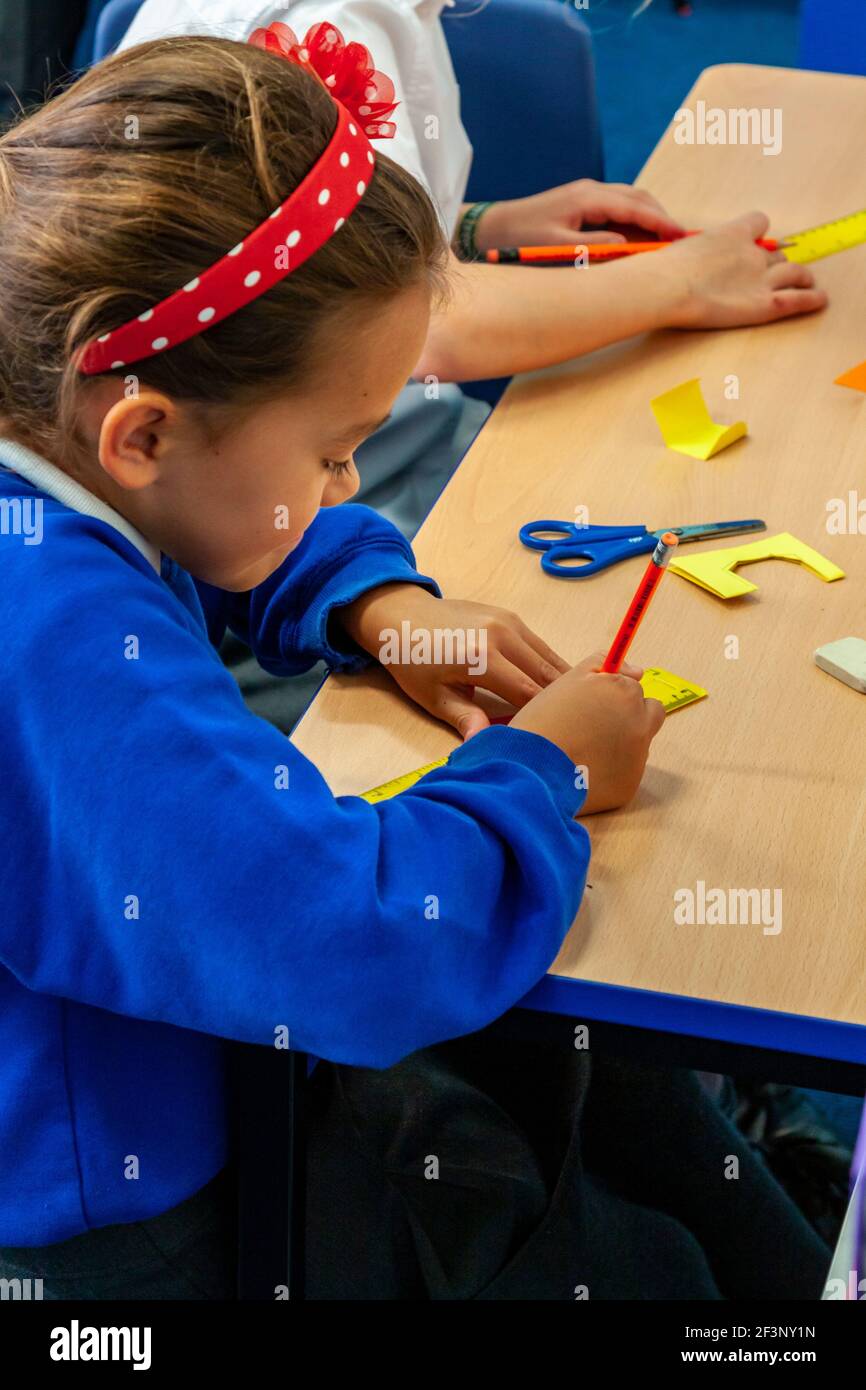 I bambini della scuola primaria scrivono e tagliano le forme dalla carta su una scrivania durante una lezione. Foto Stock