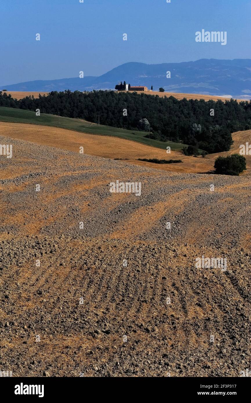 La stoppia grigia di argilla e arancio di un campo di grano arato nelle Crete Senesi contrasta con boschi, praterie e lontane colline blu in questa vista della Cappella della Madonna di Vitaleta nella Val d’Orcia, Toscana, Italia. La cappella, fondata qualche tempo prima del 1590, è un simbolo iconico del Rinascimento italiano, patrimonio dell'umanità dell'UNESCO. Foto Stock