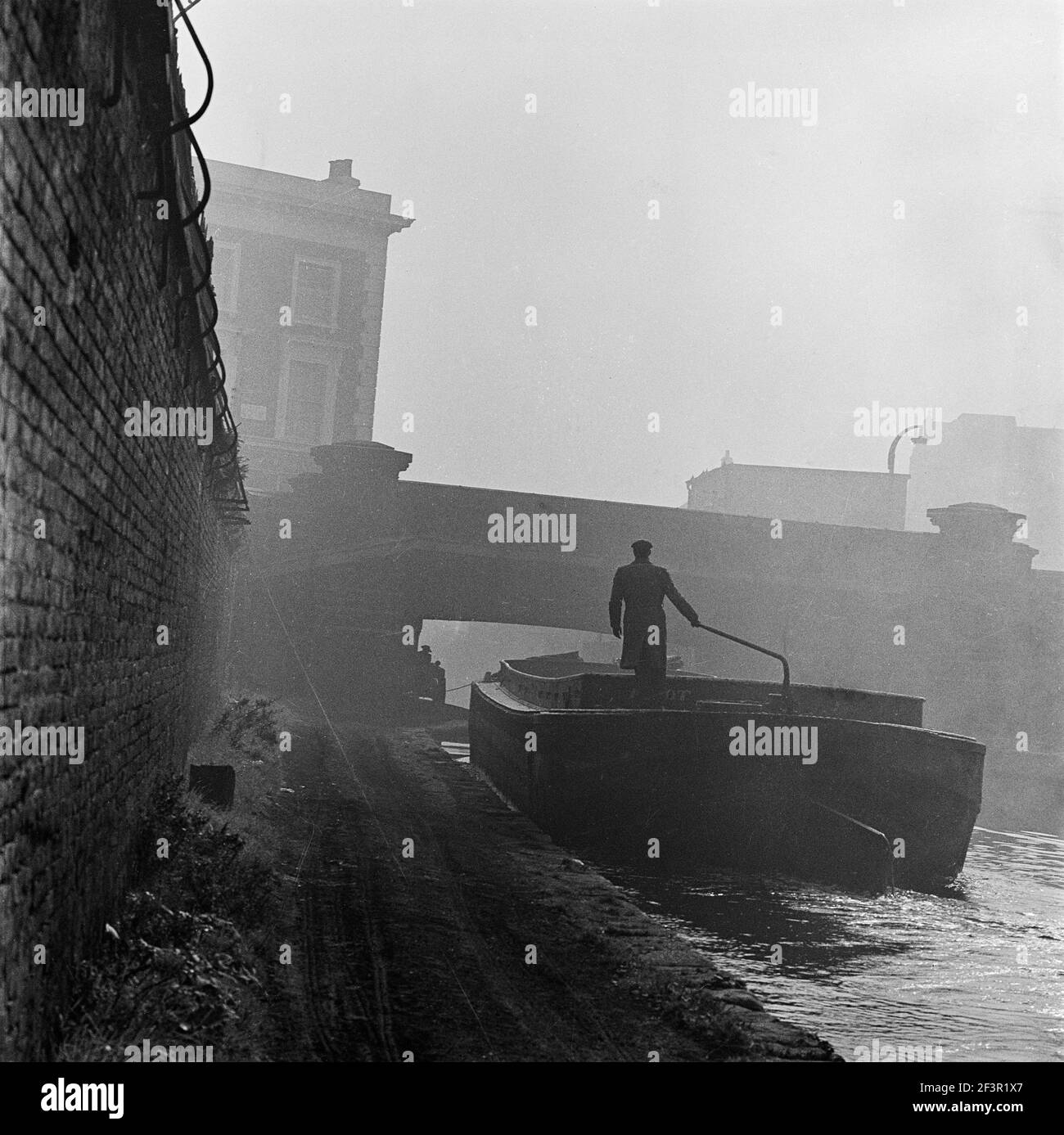 GRAND UNION CANAL, LONDRA. Una vista da un torreggiante canale nebbiosa che mostra un uomo che guida una chiatta di canale industriale lungo il canale verso un ponte stradale. Foto Stock