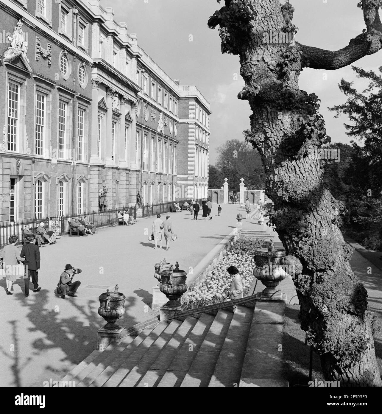 HAMPTON COURT PALACE, Greater London. Le persone che passeggiano fuori dal fronte sud per l'Hampton Court Palace con un uomo accovacciato che scatta una fotografia di un w Foto Stock