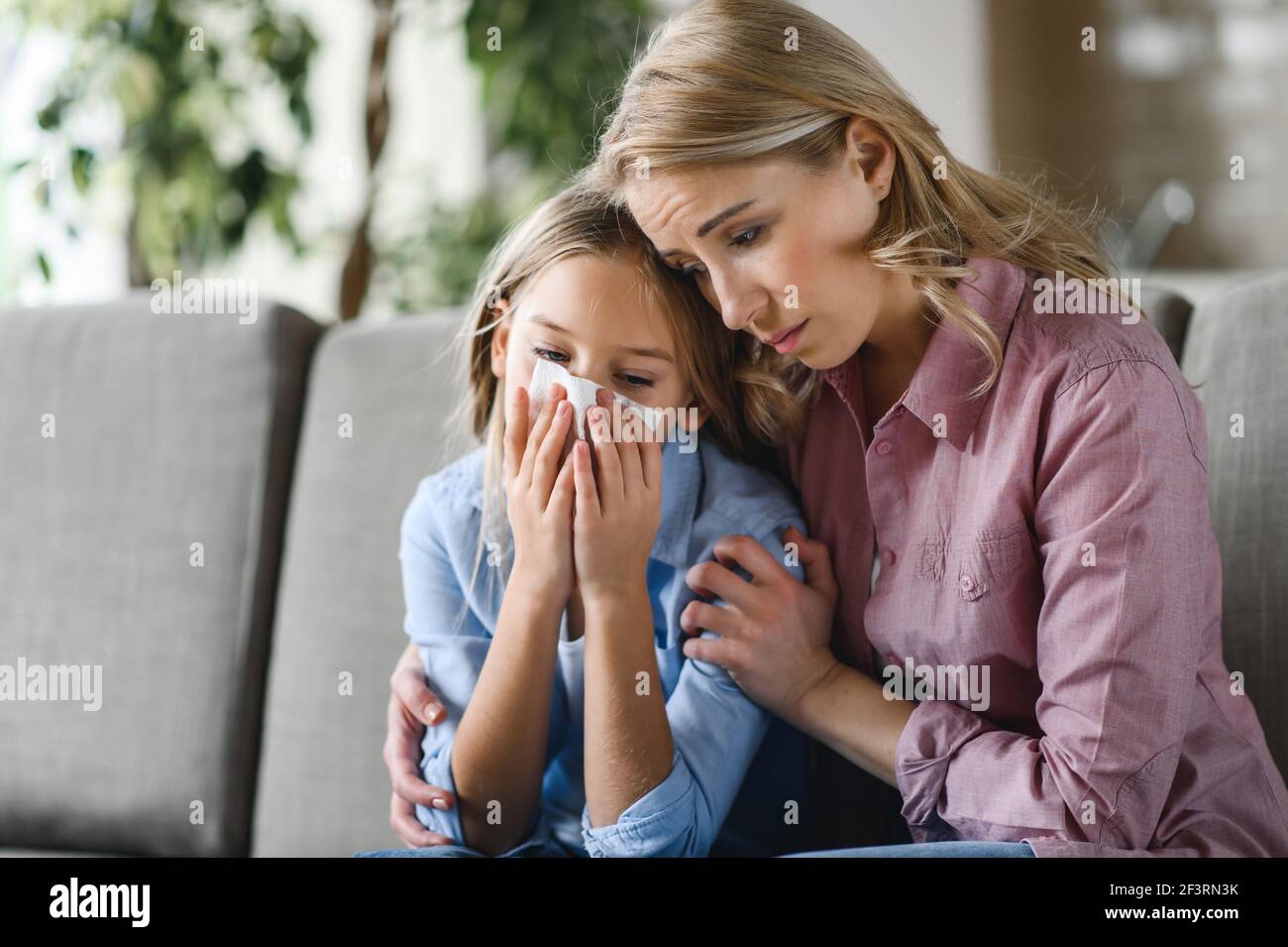 Bambina malsana e sua madre seduta a casa sul divano. La giovane mamma bionda caucasica abbraccia ansiosamente sua figlia, sostenendola perché è malata, si preoccupa di sua figlia Foto Stock