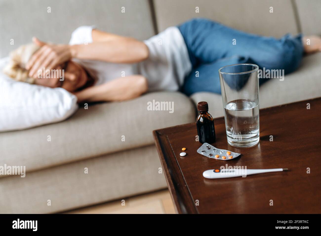 Donna adulta malata defocata a casa sul divano. Una donna malata caucasica di mezza età ha mal di testa, stringendo la testa, un bicchiere d'acqua sul tavolo e un termometro con pillole, hanno bisogno di un trattamento Foto Stock