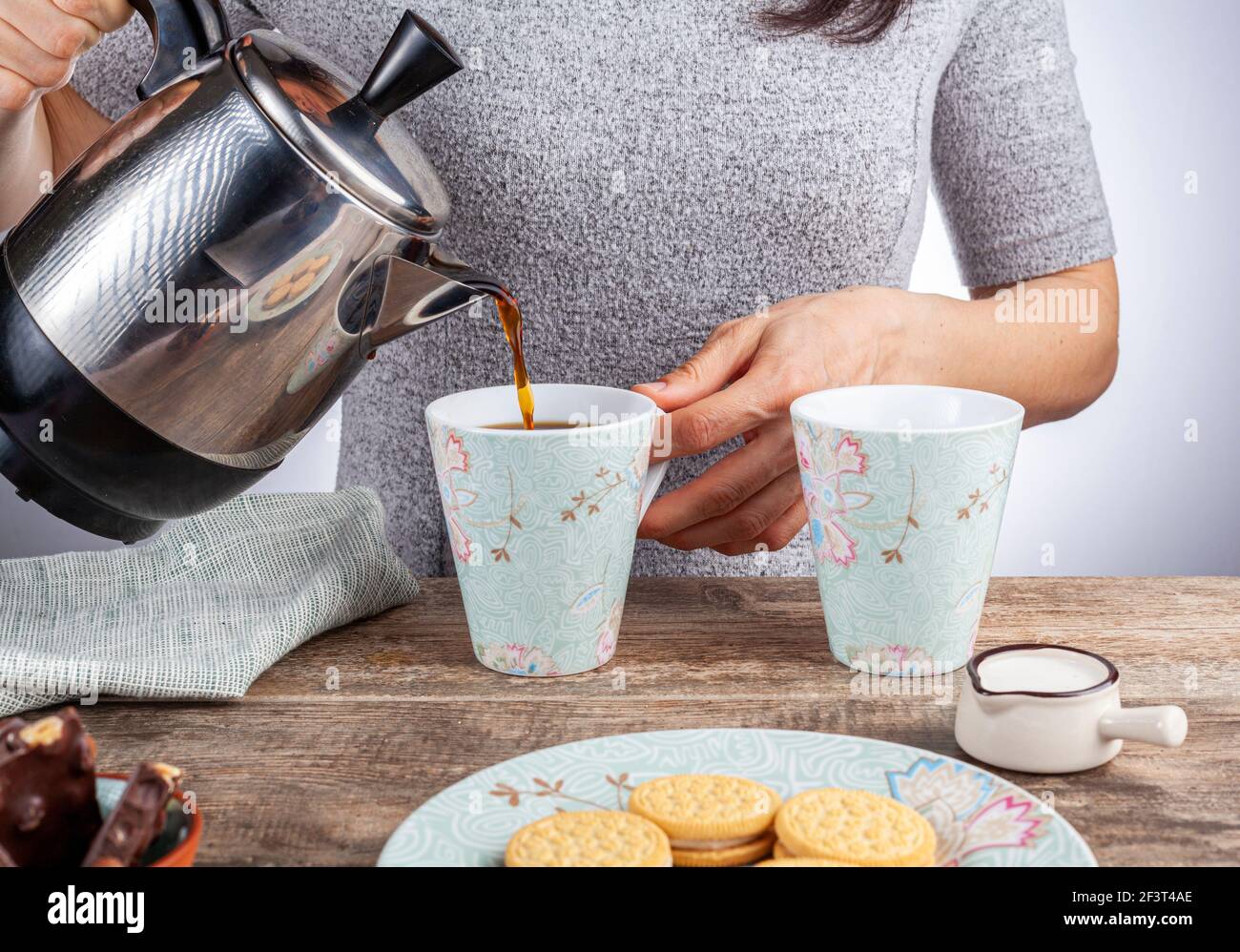 un concetto di tempo del tè o del caffè con biscotti sandwich, barrette di tazza di ceramica estetica al cioccolato e piatti, nonché una mini caraffa crematrice su legnoso Foto Stock