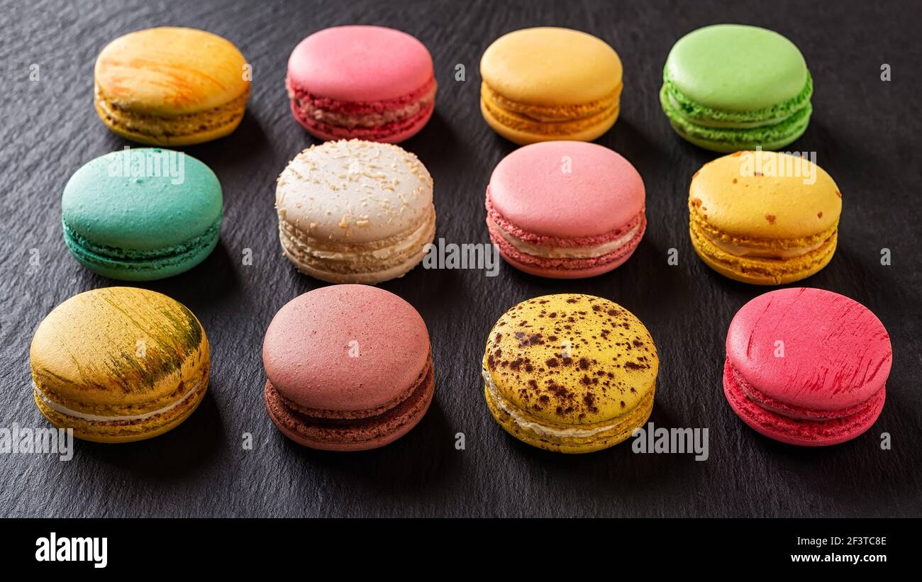 Set di dodici macaroni multicolore su sfondo nero. Dozzina di varietà di torte di macaron colorate. Delizioso dessert francese. Dolci di farina di mandorle Foto Stock