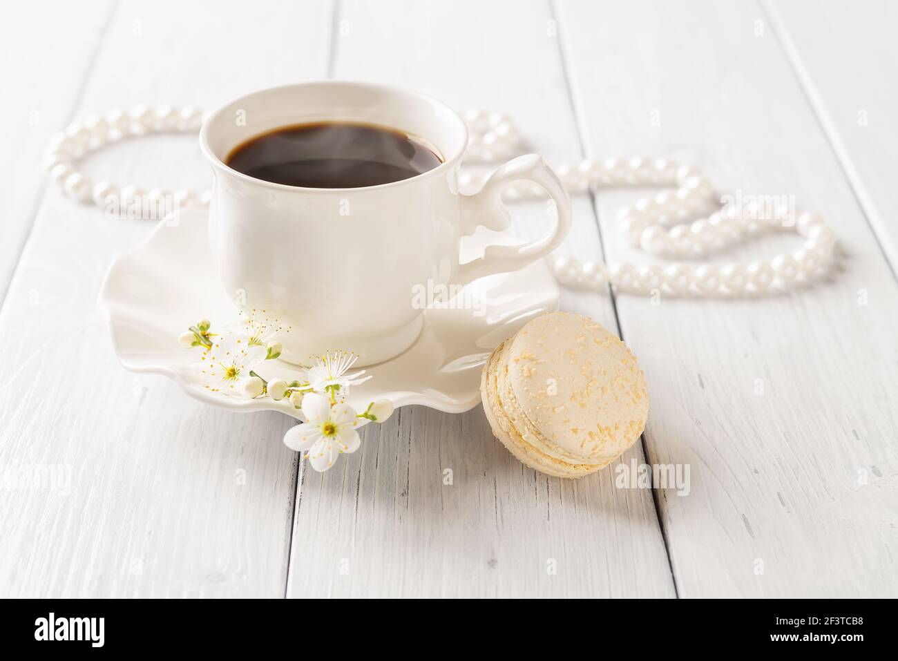 Un macaroon beige e una tazza di caffè caldo contro le perle su un tavolo di legno bianco. Colazione dolce con delizioso dessert francese. San Valentino. Foto Stock