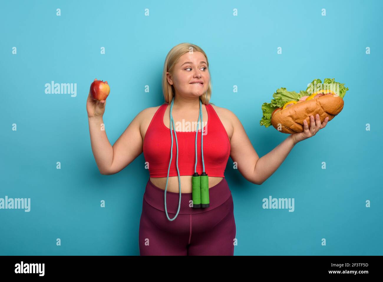 Fat girl è indecisa a mangiare una mela o un grande sandwich. Sfondo ciano Foto Stock