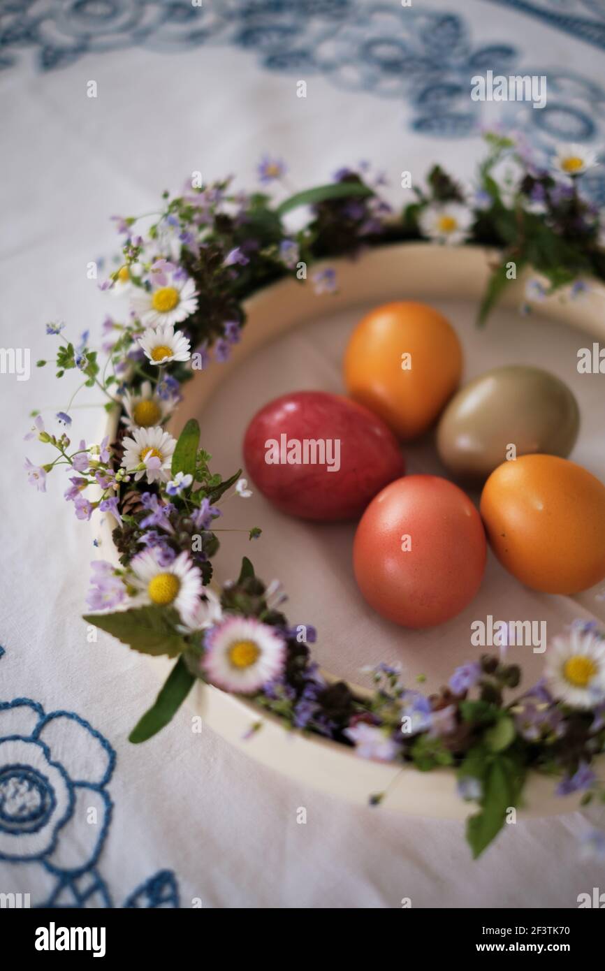 Le uova di Pasqua di colore arancio e rossastro si trovano al centro di un'antica corona di porcellana ripiena di fresco margherite su un panno da tavolo Foto Stock