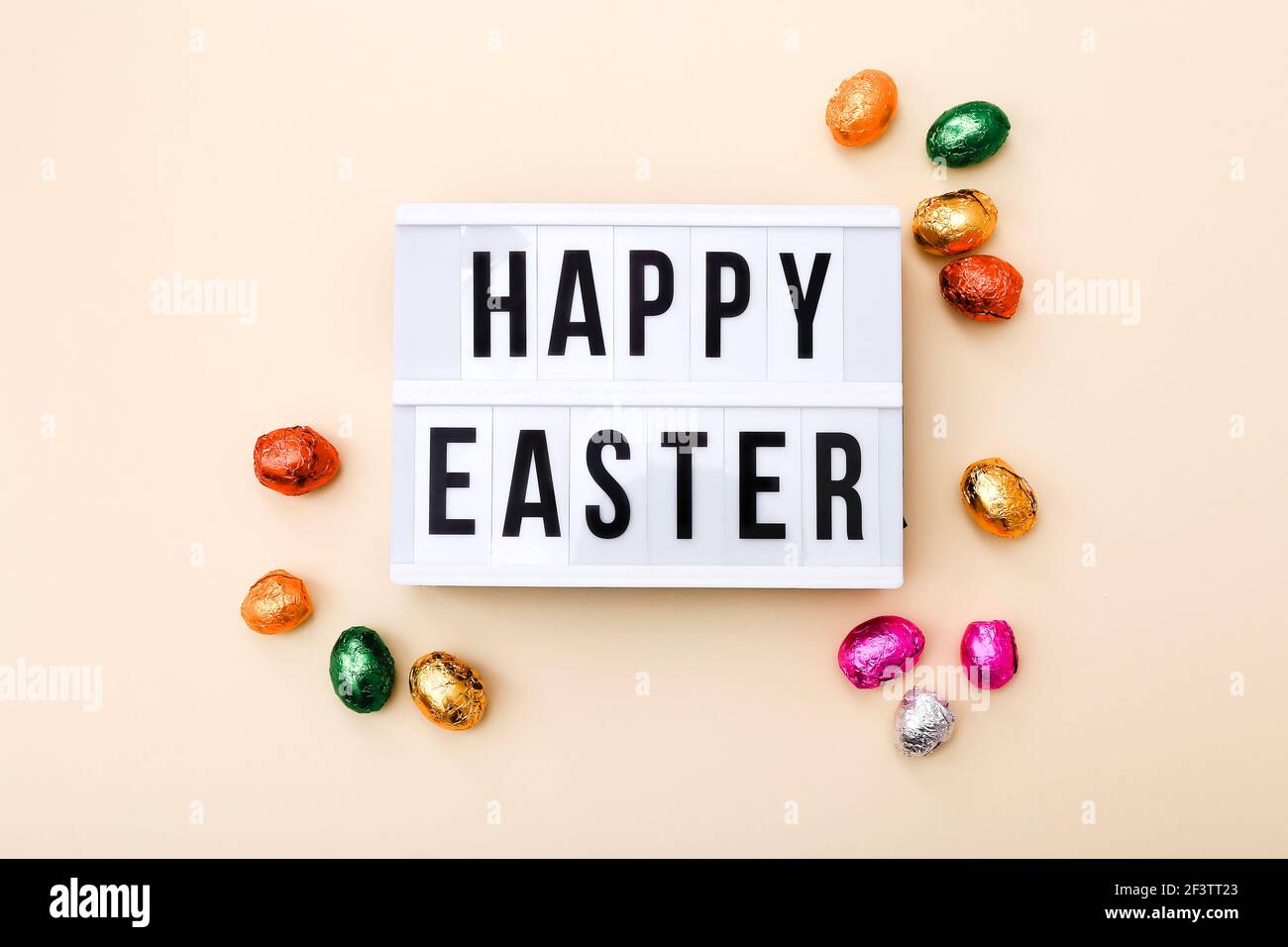 Auguri di Pasqua sulla scatola luminosa con mini uova di cioccolato avvolte in un foglio colorato su uno sfondo beige, vista dall'alto Foto Stock