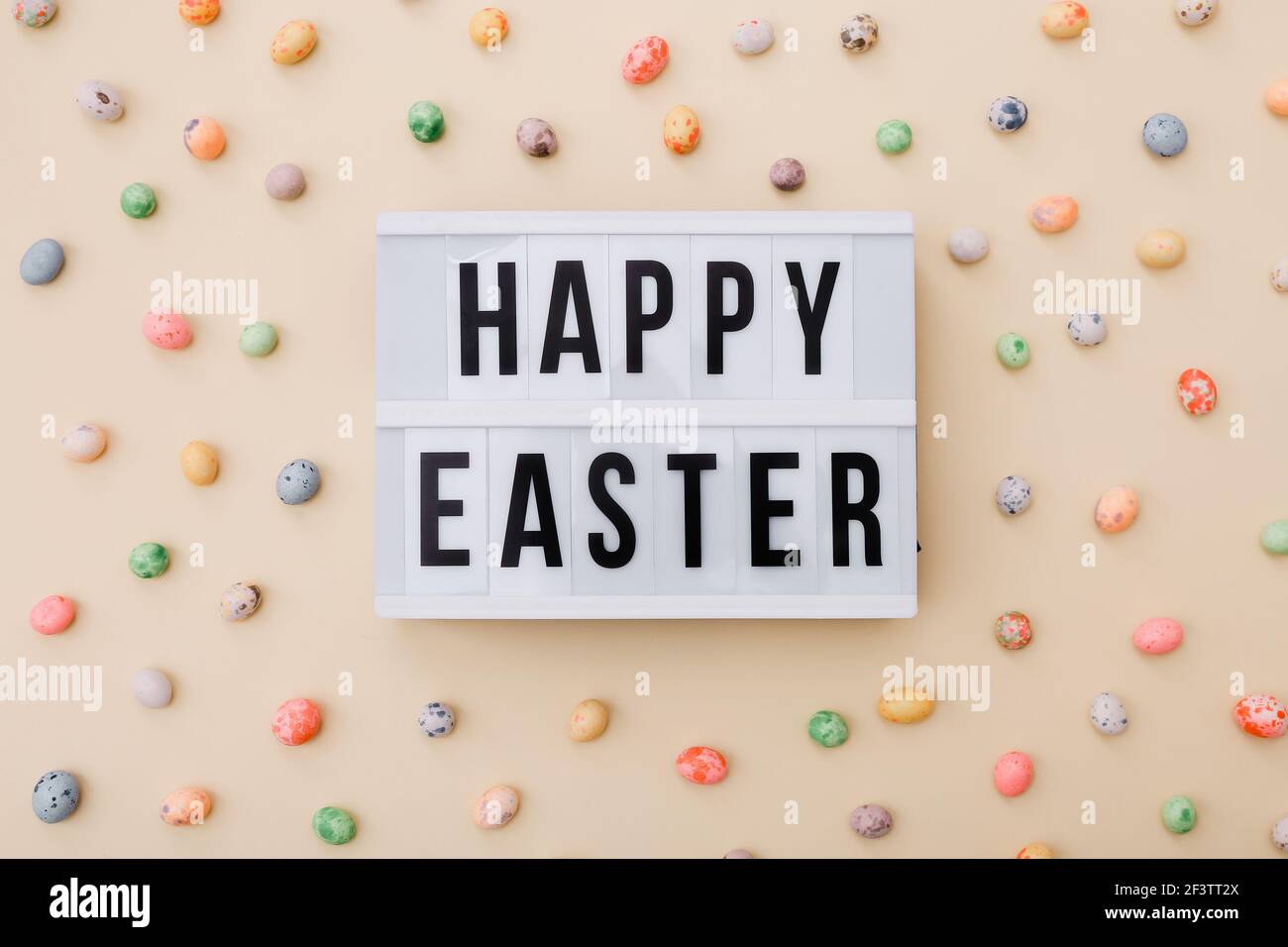 Auguri di Pasqua sulla scatola luminosa con mini uova di cioccolato sparse su uno sfondo beige, vista dall'alto Foto Stock