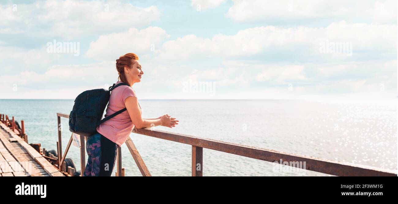 Banner. Una donna adulta felice in abbigliamento sportivo con uno zaino in posa su un molo. Sullo sfondo, il mare e il cielo. Spazio di copia. Concetto di stile di vita attivo Foto Stock