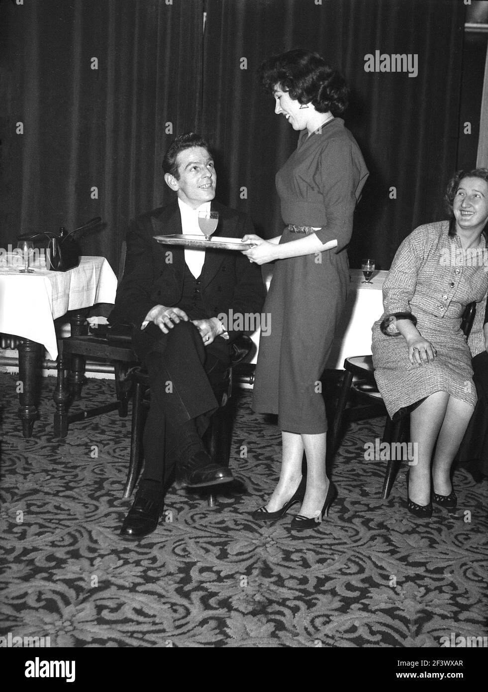1957, storica, all'interno di una sala funzioni di un hotel, una giovane donna attraente che porta un vassoio con un drink per un uomo in una giacca da pranzo, forse il leader della band, al divertimento di una ospite femminile seduto vicino, Victoria Hotel, Leeds, Inghilterra, Regno Unito. Foto Stock