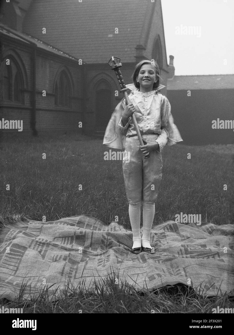 1958, storica, una giovane ragazza in costume, in possesso di un bastone, in piedi su un tappeto nel terreno di una chiesa per una foto, prima di partecipare alla tradizionale sfilata e festival del giorno di maggio, Leeds, Inghilterra, Regno Unito. Foto Stock