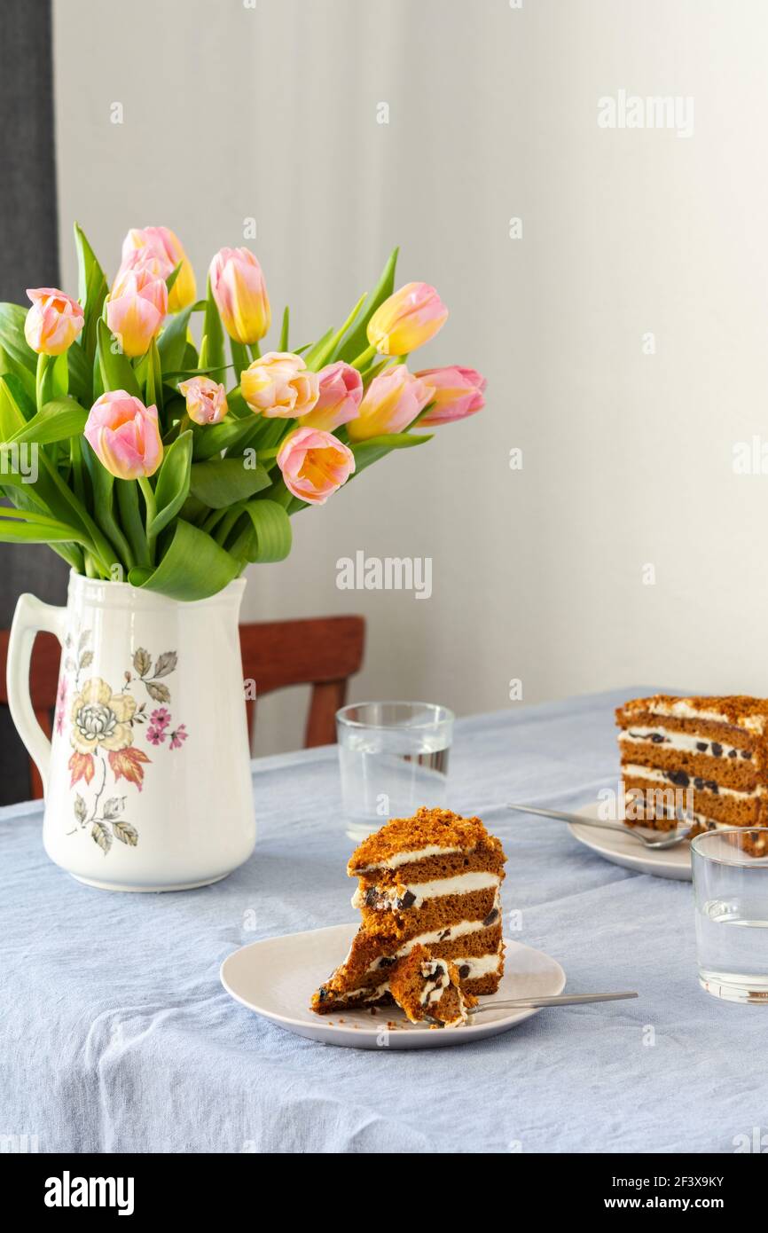 Bel bouquet primaverile di tulipani rosa in una brocca, porzioni di torta con prugne su piatti e bicchieri d'acqua su un tavolo con una tovaglia blu Foto Stock