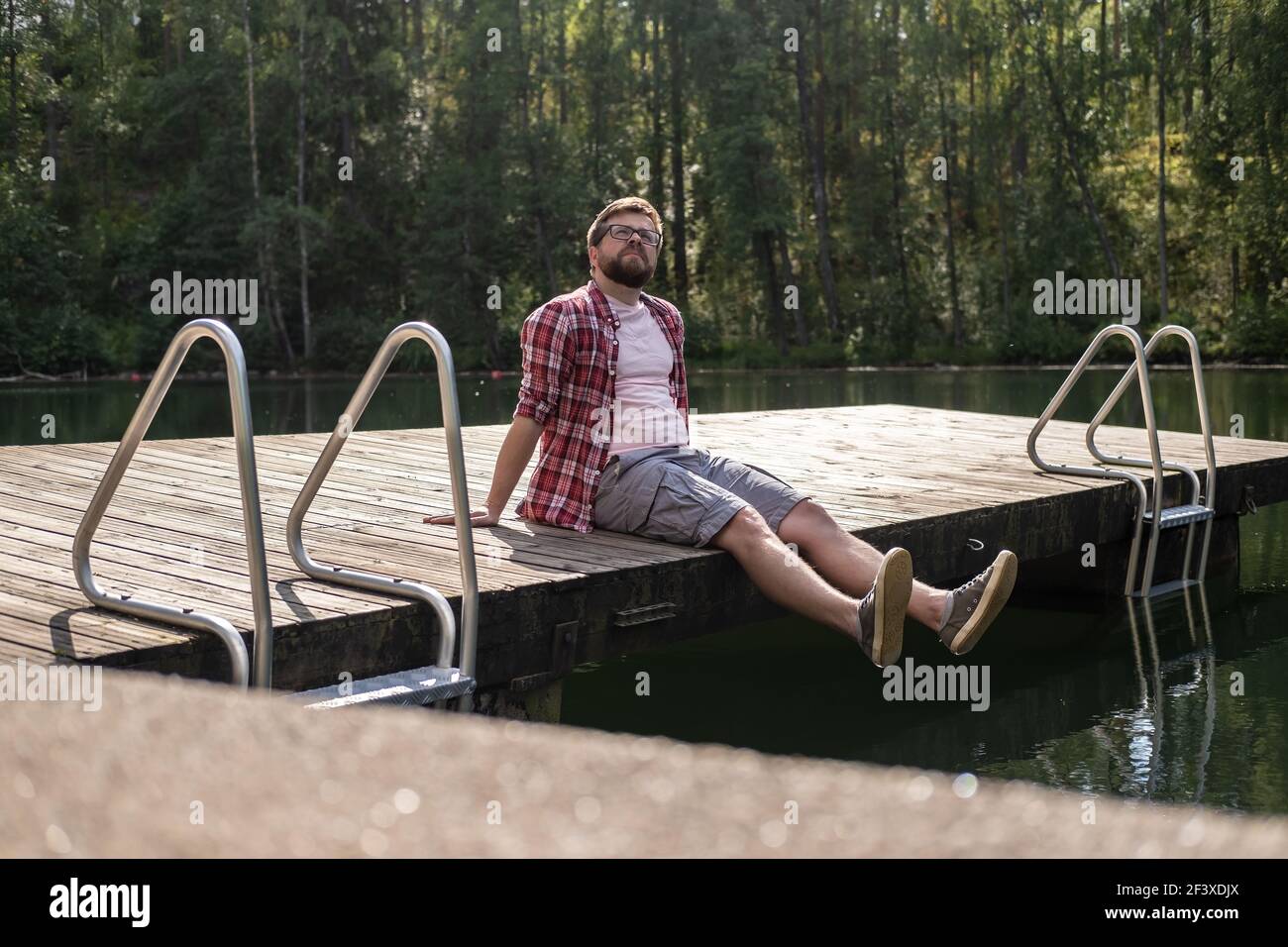 L'uomo calmo siede sul bordo di un molo di legno e guarda in su pensieroso, sullo sfondo di un lago di foresta calmo. Stile di vita. Foto Stock