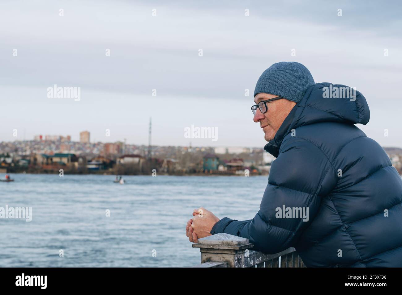 Ritratto in primo piano dell'uomo di mezza età in bicchieri in piedi sull'argine del fiume e guardando in lontananza. Un uomo in una giacca calda e cappello. Foto Stock