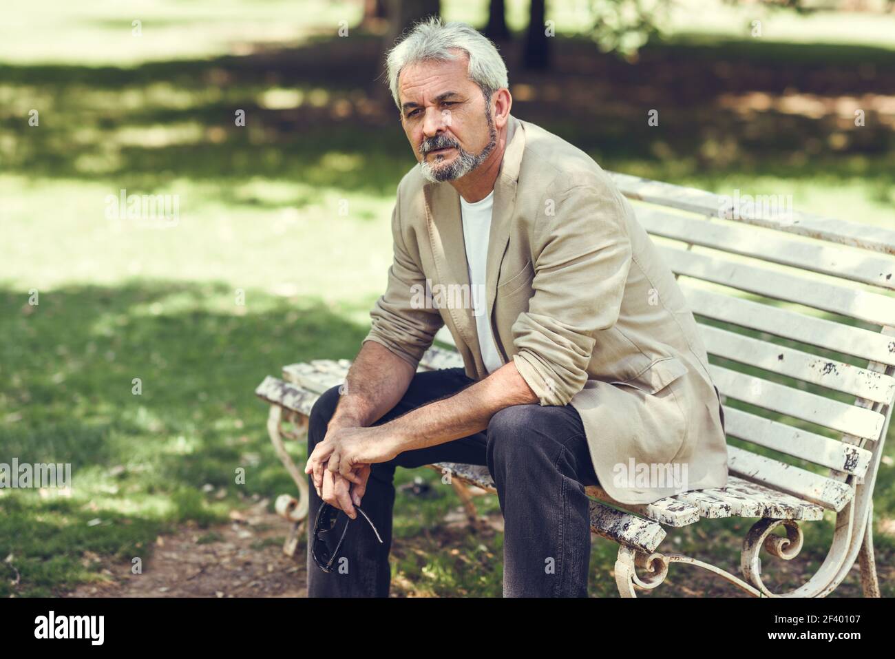 Uomo maturo pensivo seduto su panchina in un parco urbano. Ritratto di un uomo maturo pensivo seduto su una panchina in un parco urbano. Uomo anziano con capelli bianchi e barba che indossa abiti casual. Foto Stock