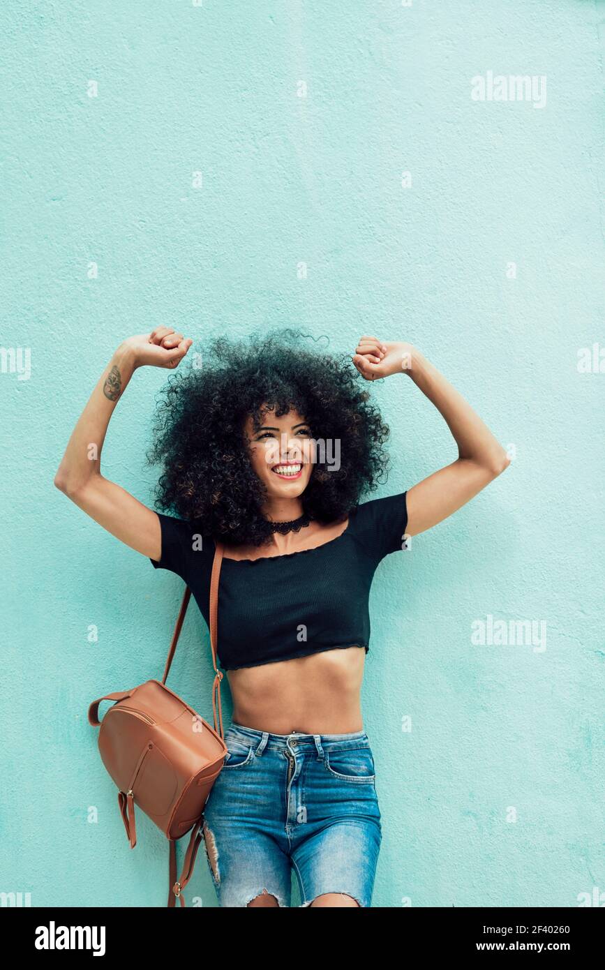 Donna nera divertente con i capelli afro che alzano le braccia all'aperto. Donna nera sorridente con capelli ricci che alzano le braccia all'aperto. Donna che indossa abiti casual su sfondo urbano. Concetto di stile di vita Foto Stock