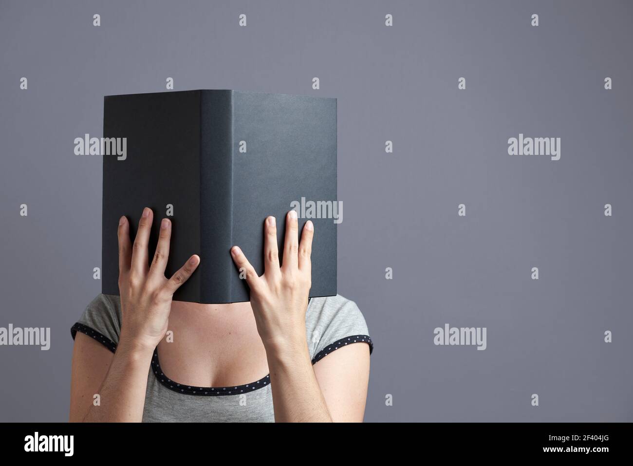 Giovane donna con il suo volto nascosto dietro un libro nero che tiene con entrambe le mani, aperto, la lettura. Verticale con sfondo grigio e spazio per la copia. Foto Stock