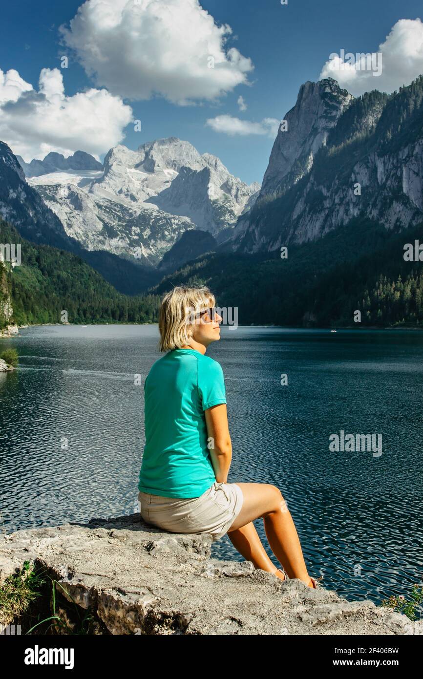 Bionda ragazza seduta sulla roccia godendo la vista delle montagne maestose e delle acque turchesi cristallo in Gosau See, Austria, ghiacciaio Dachstein in background. Foto Stock