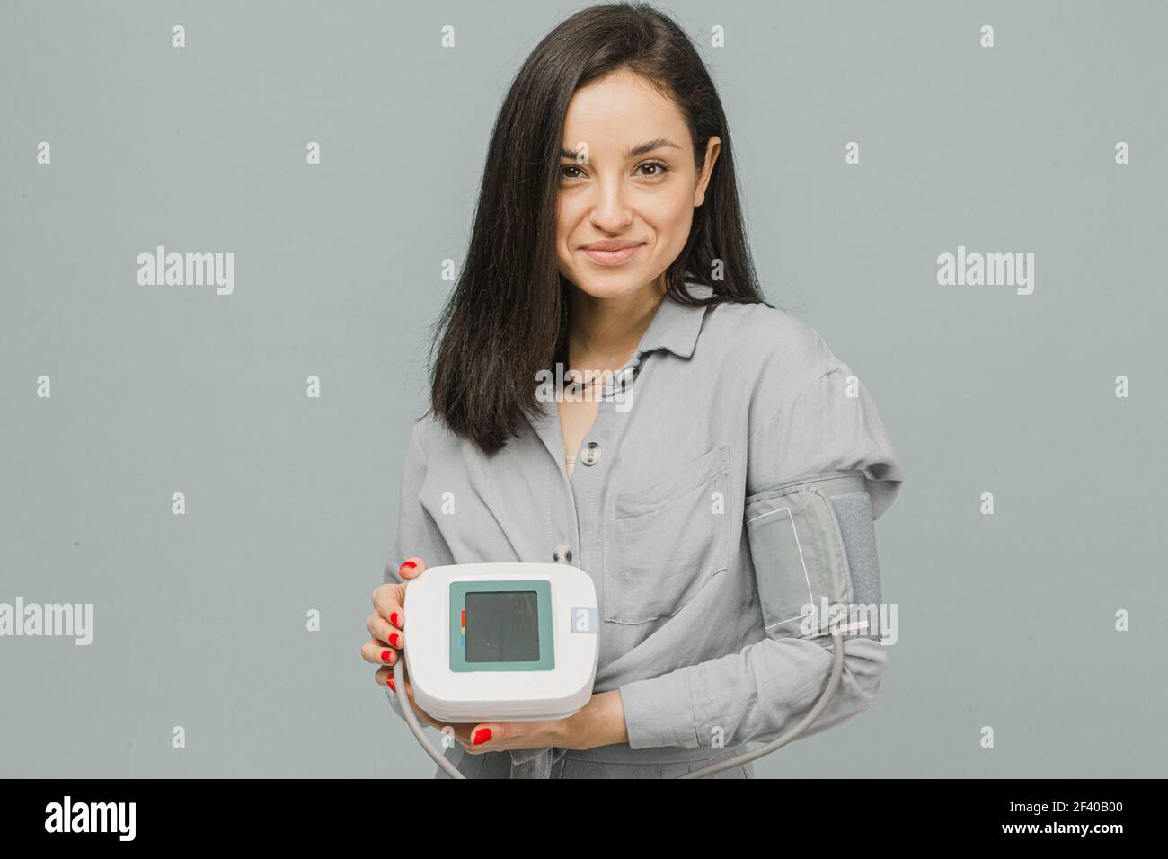 Foto di cute femmina tiene manometro, controlla la sua salute del cuore. Indossa un abito grigio, isolato sfondo di colore grigio Foto Stock