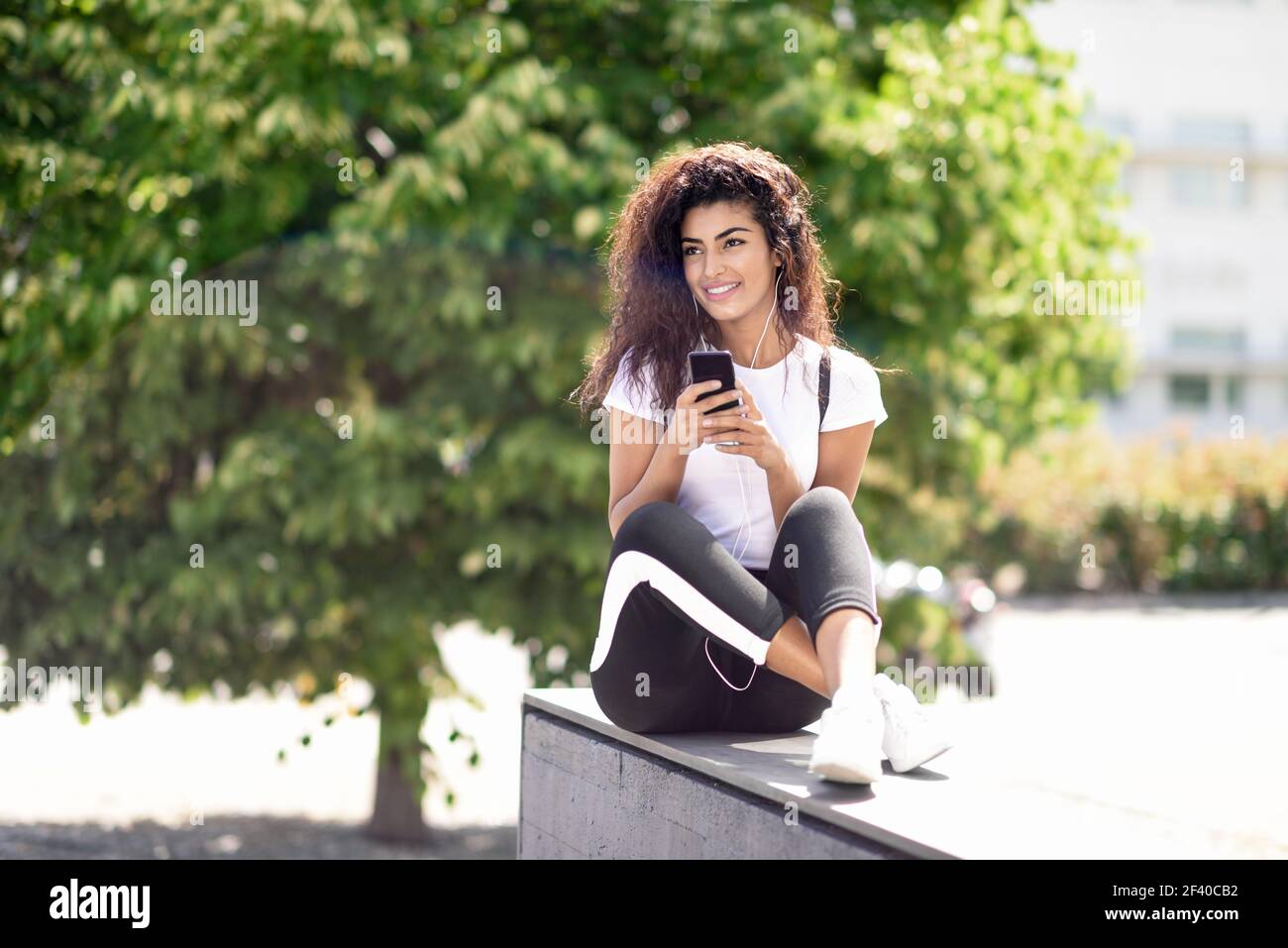Moderna donna araba ascoltando la musica con gli auricolari all'esterno. Nord ragazza africana in sportswear con ricci seduta di acconciatura in background urbano. Foto Stock