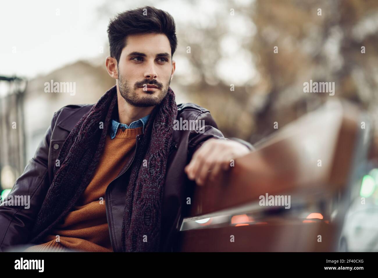 Considerato giovane uomo seduto su una panchina urbana. Ragazzo attraente con moderni acconciatura con sguardo perso per strada. Foto Stock