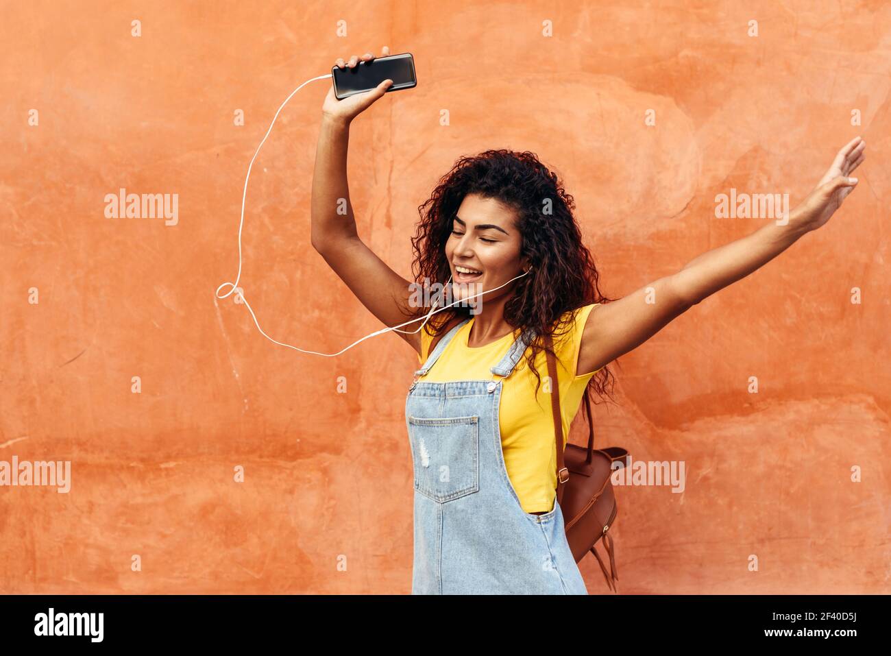 Felice ragazza arabo ascoltare musica e ballare con gli auricolari all'esterno. Donna africana in abiti casual con capelli ricci in background urbano. Foto Stock