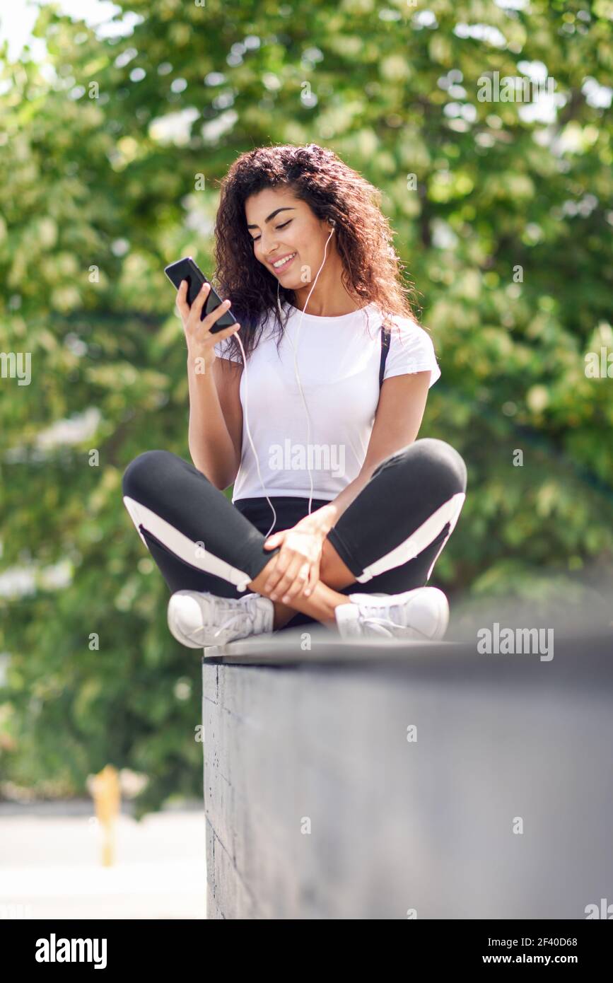 Bella donna africana che ascolta musica con auricolari e smartphone all'aperto. Ragazza araba in abiti sportivi con acconciatura riccia su sfondo urbano. Foto Stock