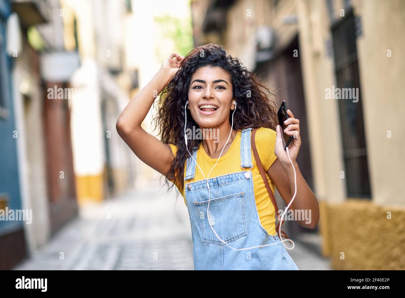 Attraente Nord donna africana ascoltando la musica con gli auricolari all'esterno. Ragazza araba in abiti casual con capelli ricci in background urbano. Foto Stock