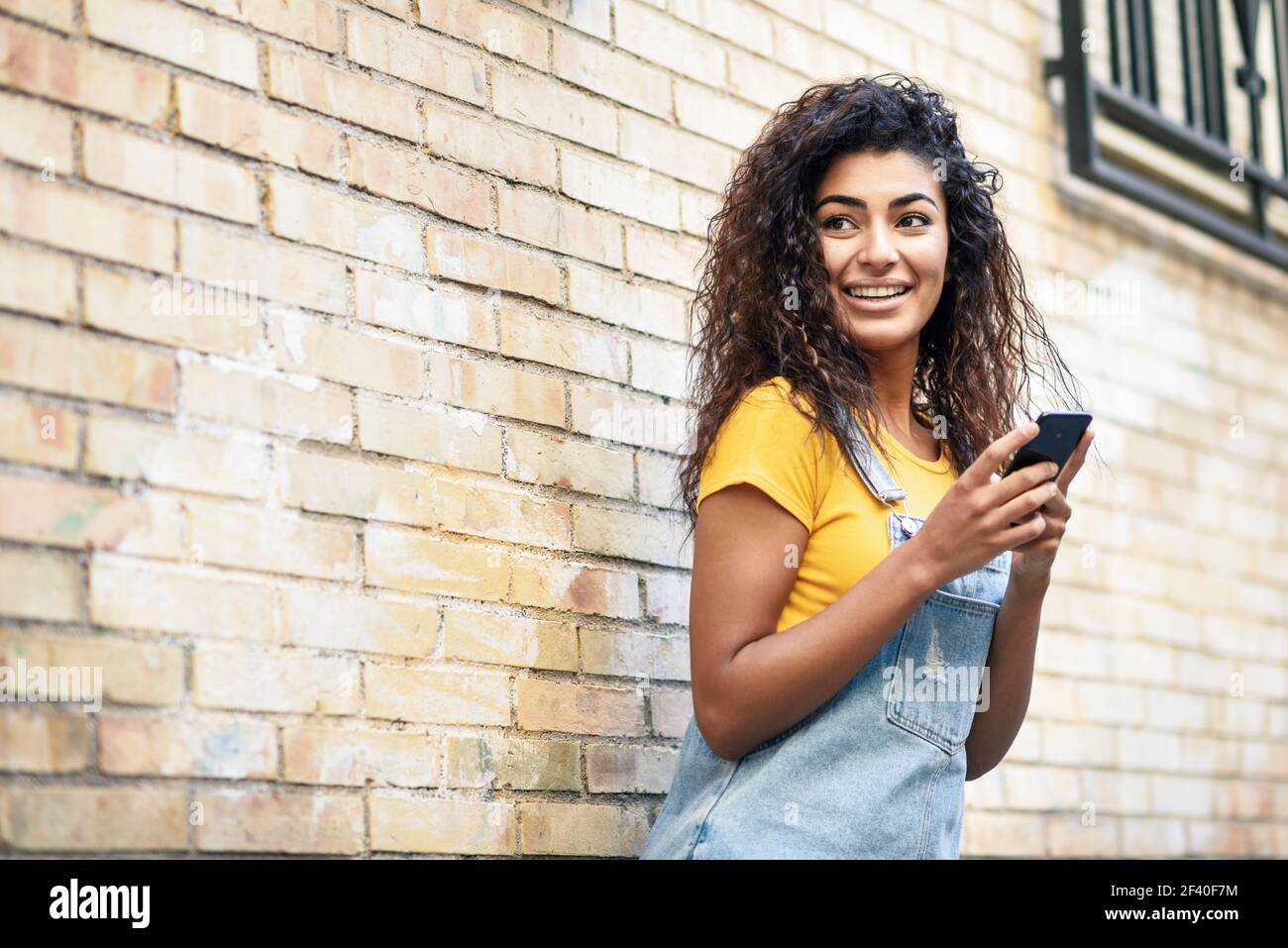 Felice ragazza araba utilizzando smart phone su un muro di mattoni. Donna sorridente con capelli ricci in abiti casual in background urbano. Foto Stock