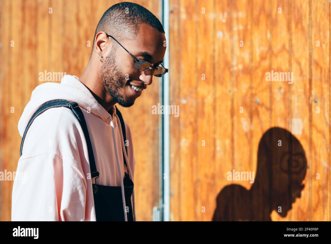 Giovane uomo nero di indossare un abbigliamento informale e occhiali da sole, sorridente contro uno sfondo di legno. Millenaria ragazzo africano con bib pants all'aperto Foto Stock