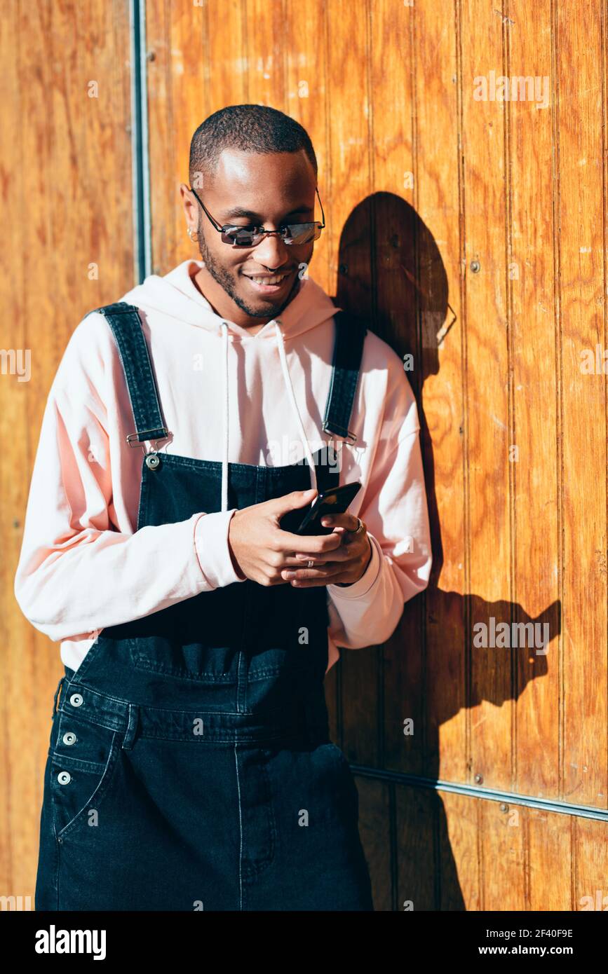 Giovane uomo nero che indossa abiti casual e occhiali da sole utilizzando smartphone su sfondo di legno. Ragazzo africano millenario con pantaloni bib all'aperto sorridente Foto Stock