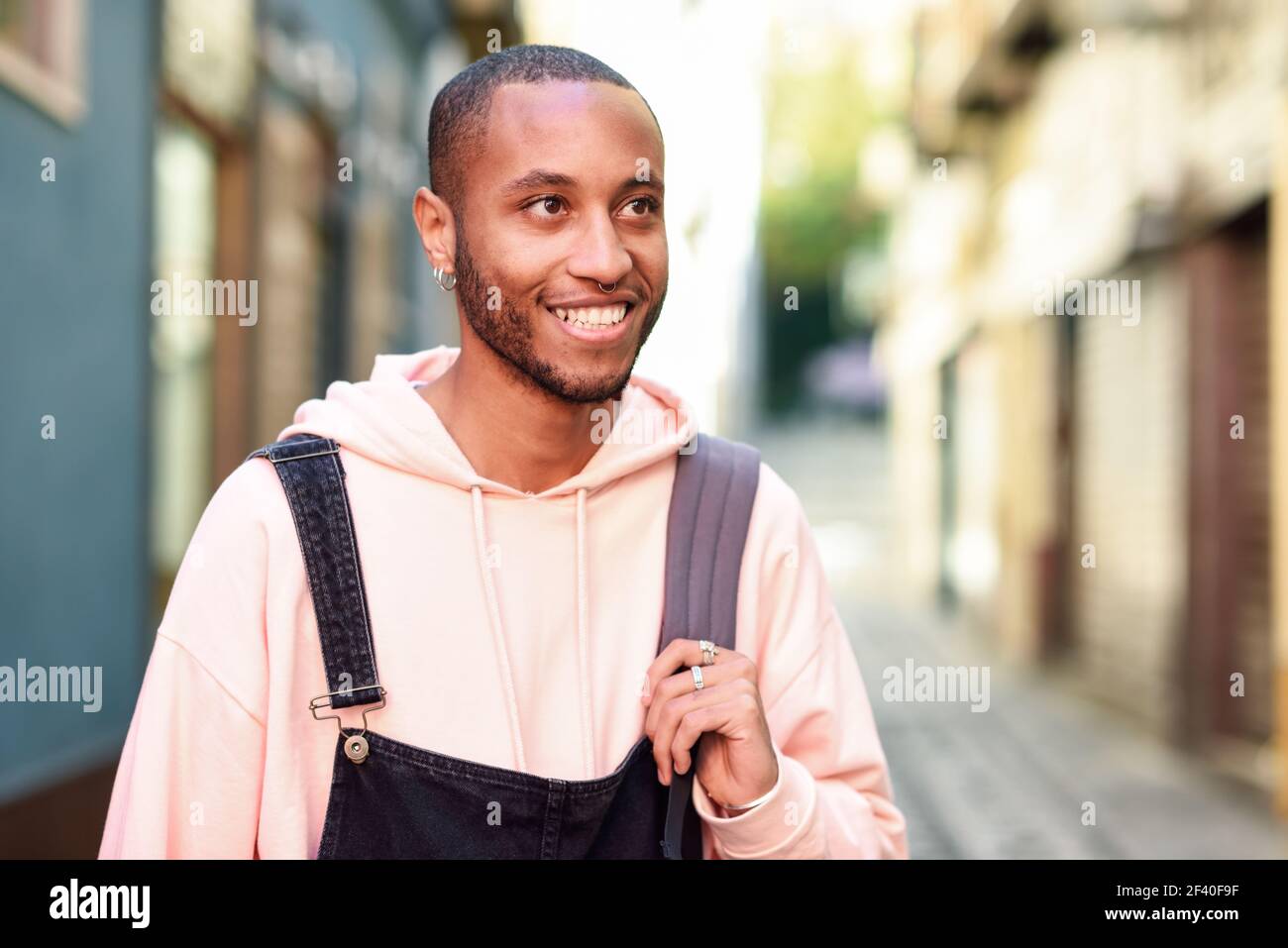 Giovane uomo nero indossando abiti casual sorridendo a piedi giù per la strada. Millenaria ragazzo africano con bib pants all'aperto Foto Stock