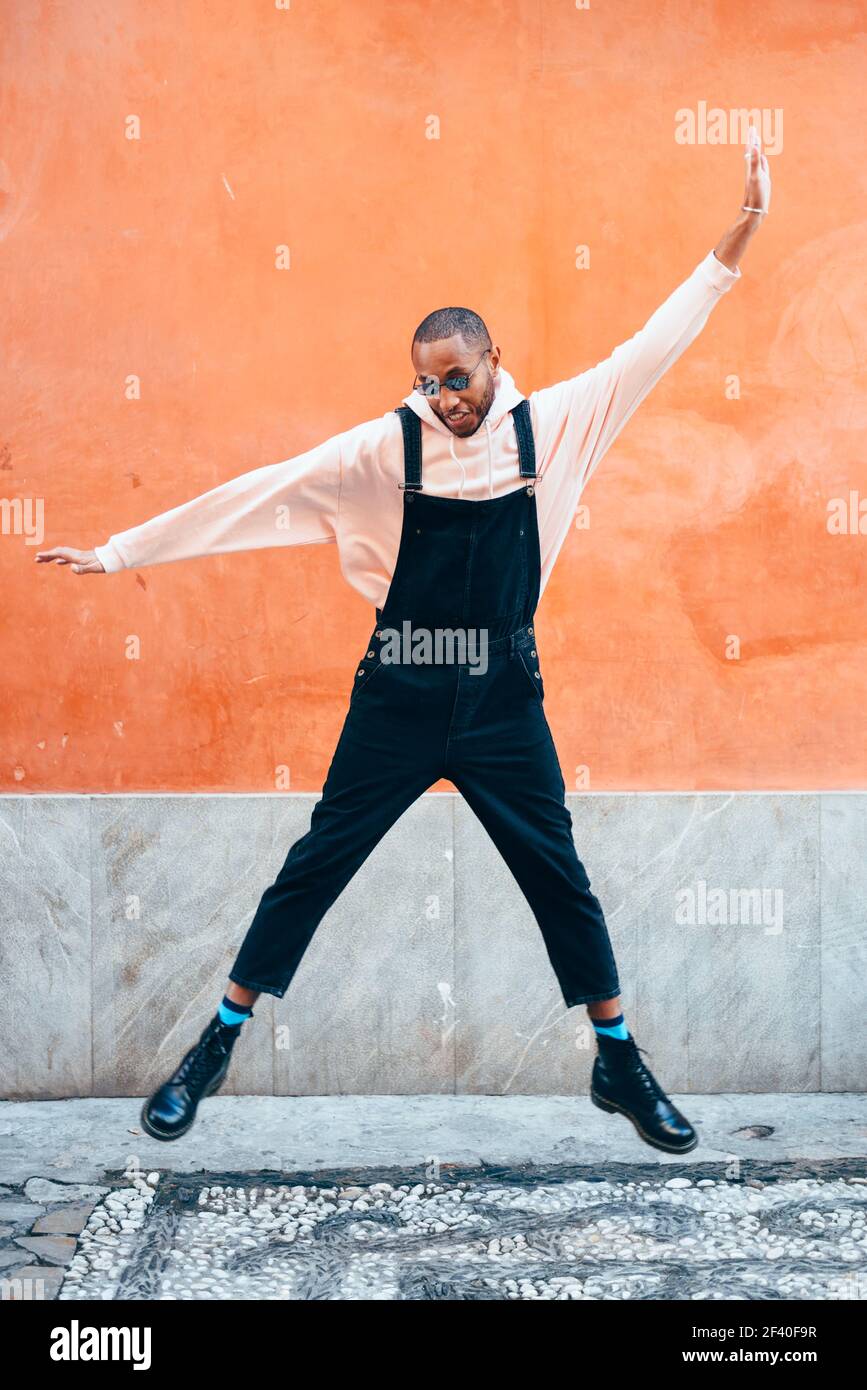 Giovane uomo nero indossando abiti casual jumping in background urbano. Il concetto di stile di vita. Millenaria ragazzo africano con bib pants all'aperto Foto Stock
