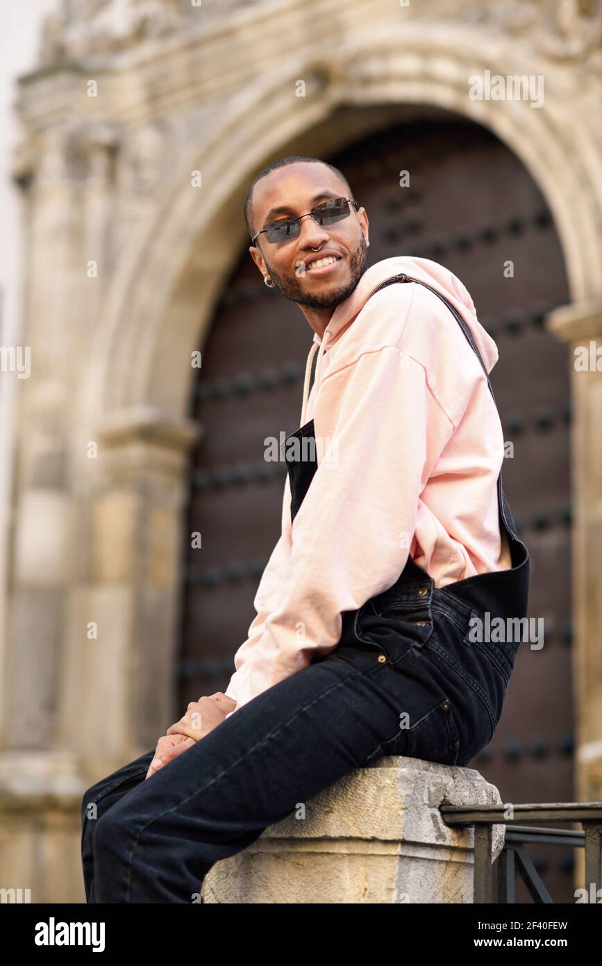 Giovane uomo nero di indossare un abbigliamento informale e occhiali da sole, seduto sul background urbano. Giovane ragazzo africano con bib pants all'aperto Foto Stock