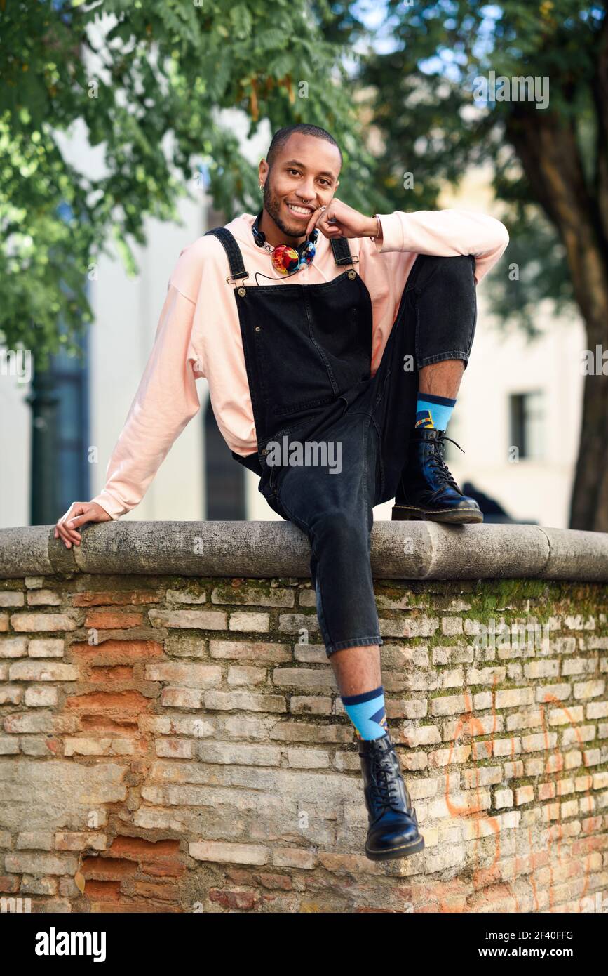 Giovane uomo nero di indossare un abbigliamento informale e cuffie seduti sul background urbano. Felice ragazzo africano con bib pants all'aperto Foto Stock