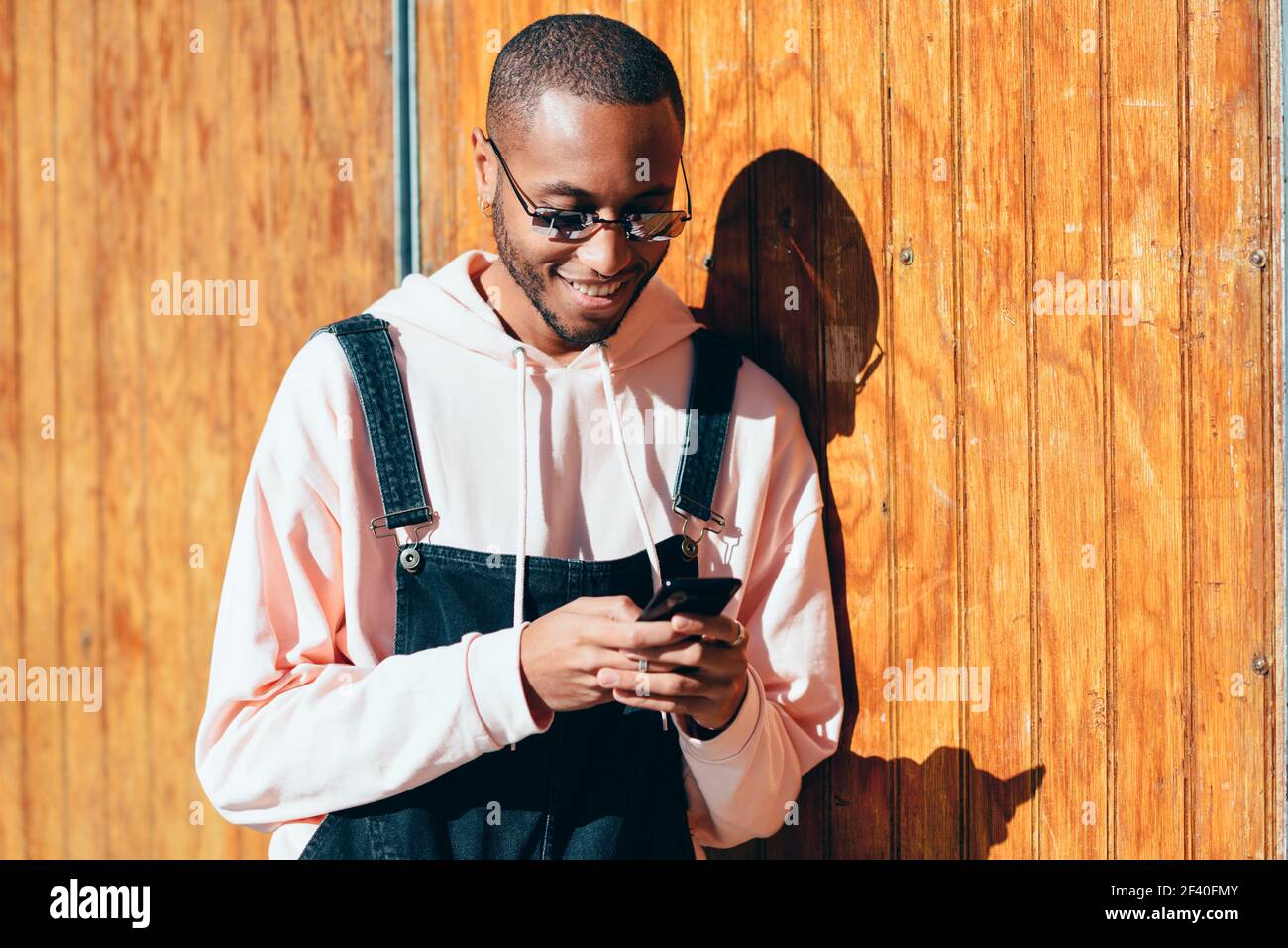 Giovane uomo nero che indossa abiti casual e occhiali da sole utilizzando smartphone su sfondo di legno. Ragazzo africano millenario con pantaloni bib all'aperto sorridente Foto Stock