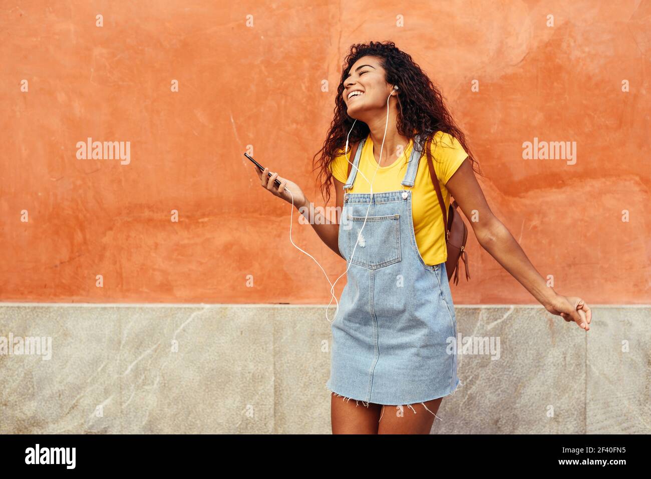 Felice donna nordafricana che ascolta musica e balla con gli auricolari all'aperto. Ragazza araba in abiti casual con capelli ricci in stile urbano. Foto Stock