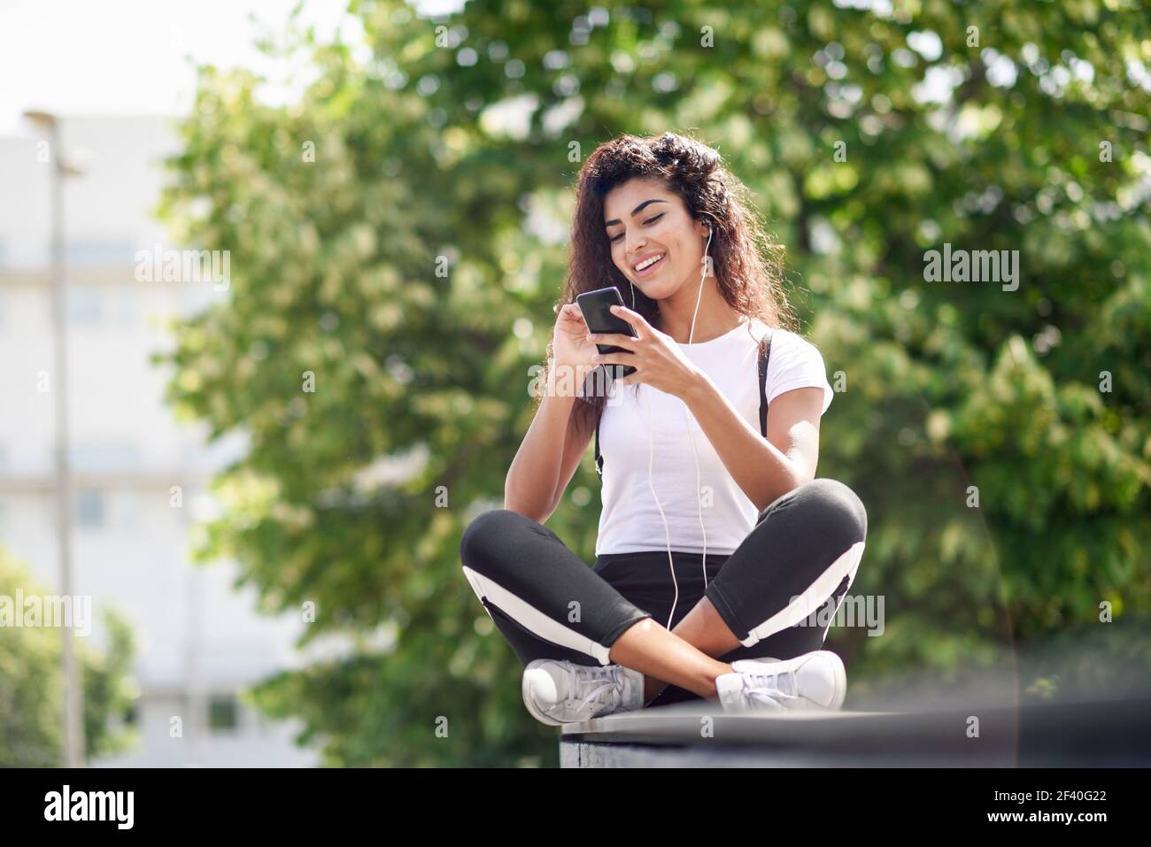 Bella donna africana che ascolta musica con auricolari e smartphone all'aperto. Ragazza araba in abiti sportivi con acconciatura riccia su sfondo urbano. Foto Stock