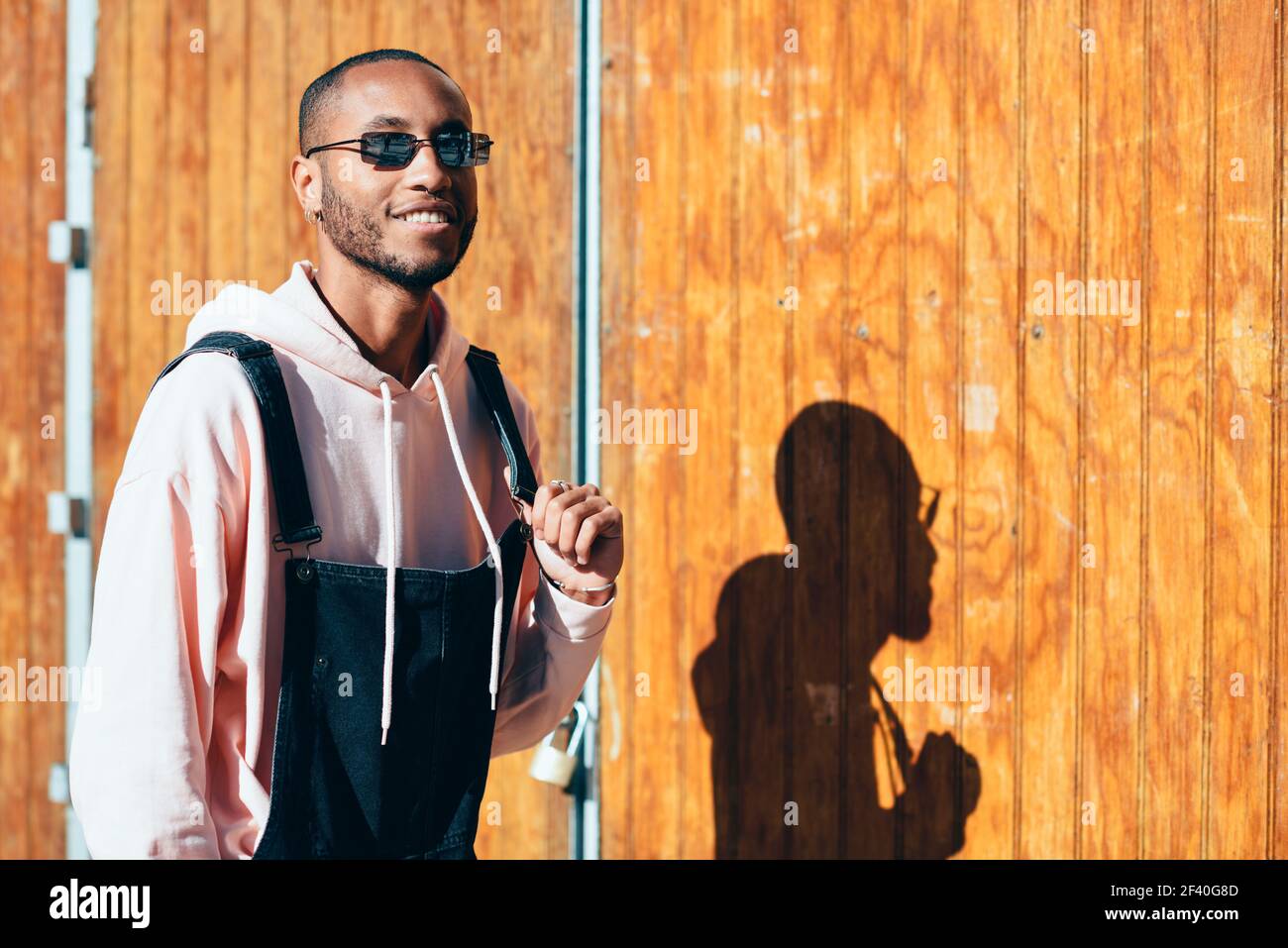 Giovane uomo nero di indossare un abbigliamento informale e occhiali da sole, sorridente contro uno sfondo di legno. Millenaria ragazzo africano con bib pants all'aperto Foto Stock