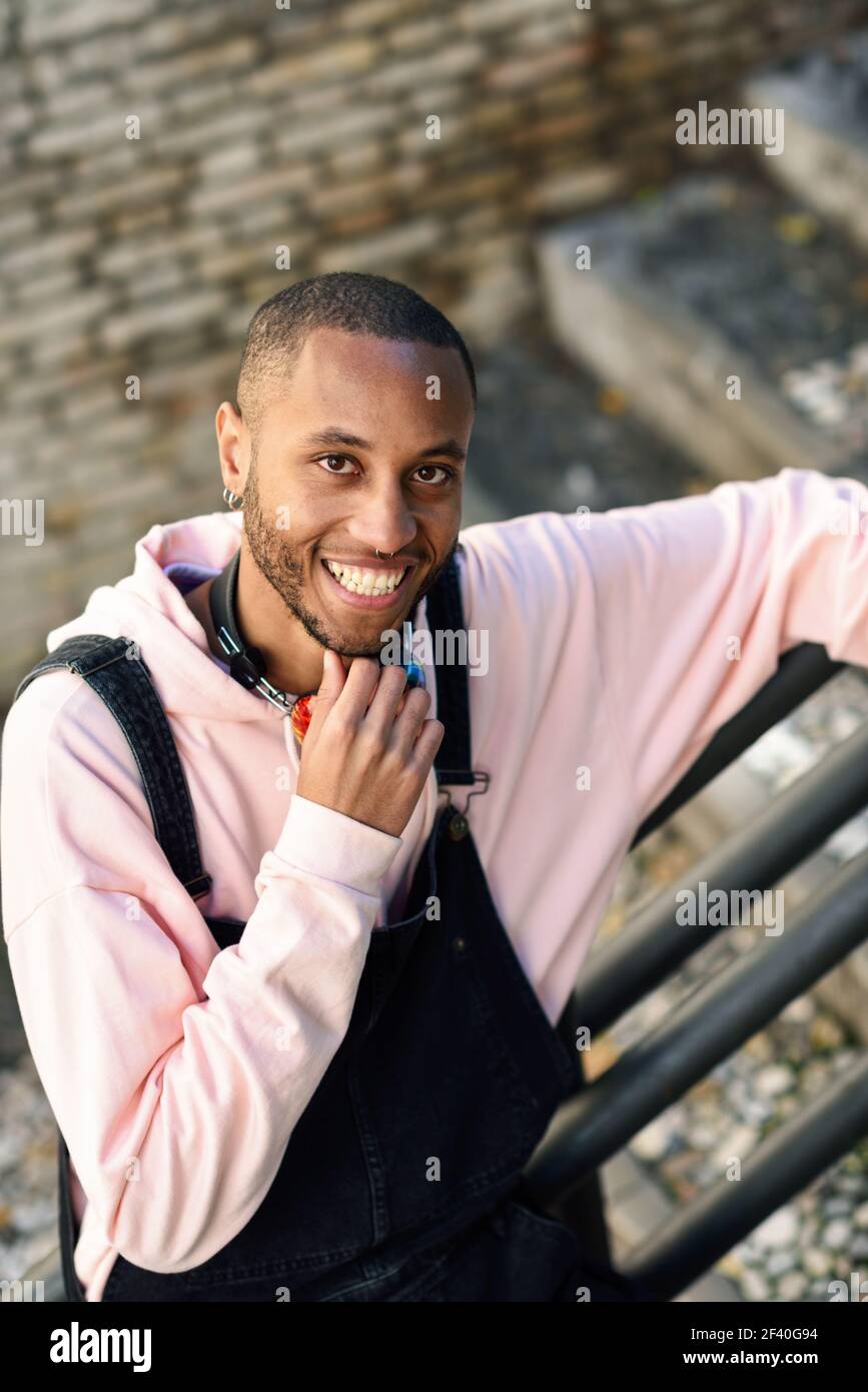 Giovane uomo nero indossando abiti casual sorridente su scala urbana. Felice ragazzo africano con bib pants all'aperto Foto Stock