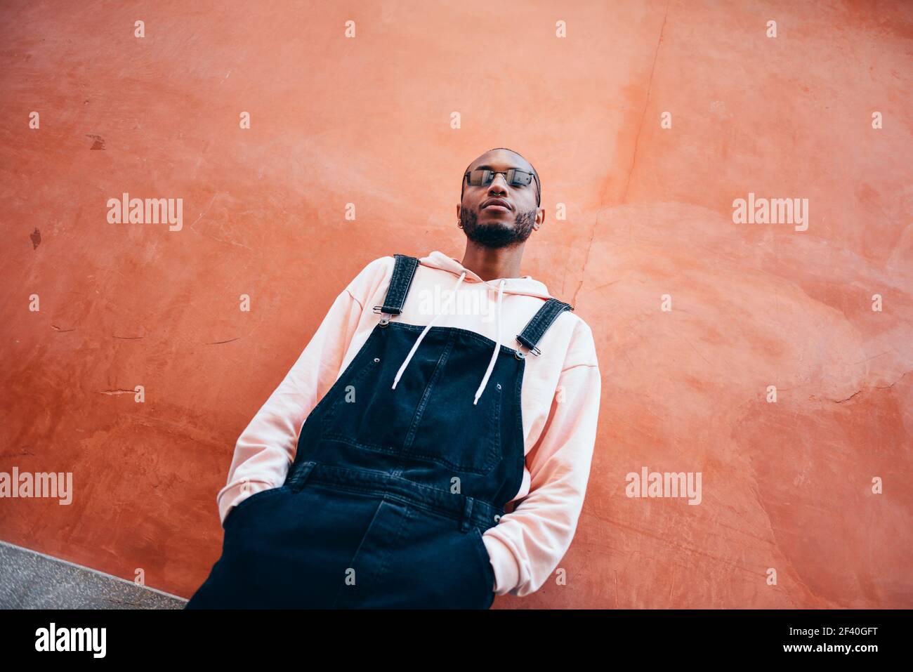 Giovane uomo nero che indossa abiti casual e occhiali da sole, su uno sfondo rosso. Ragazzo africano con pantaloni di bib all'aperto. Ragazzo africano millenario con pantaloni bib all'aperto Foto Stock