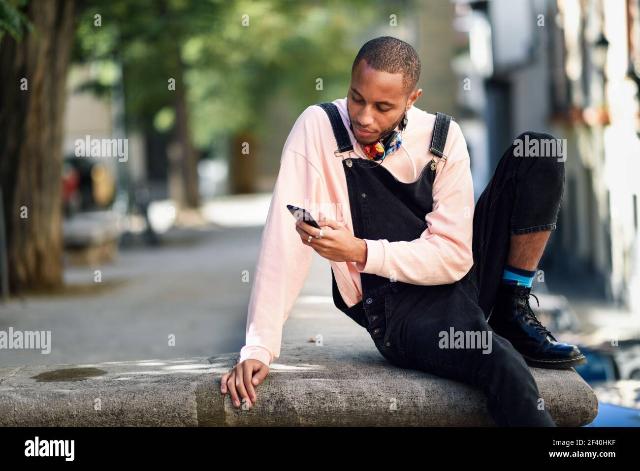 Giovane uomo nero con le cuffie seduto in strada urbana guardando il suo smartphone. Concetto di stile di vita.. Giovane uomo nero che usa lo smartphone all'aperto Foto Stock