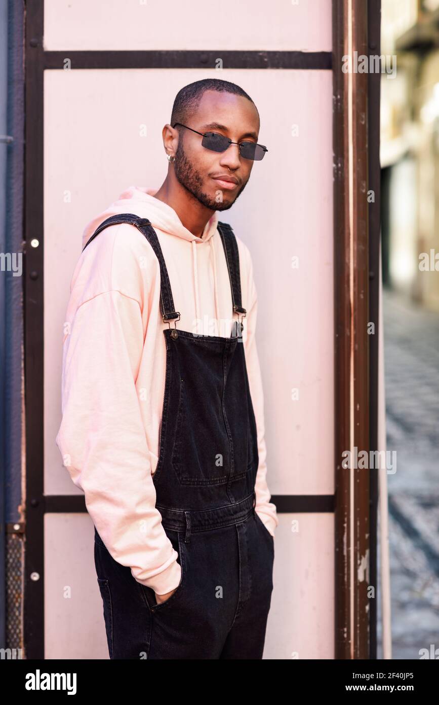 Giovane uomo nero che indossa abiti e occhiali da sole casual, su uno sfondo urbano rosa. Ragazzo africano con pantaloni di bib in piedi all'aperto. Ragazzo africano con pantaloni di bib in piedi all'aperto Foto Stock