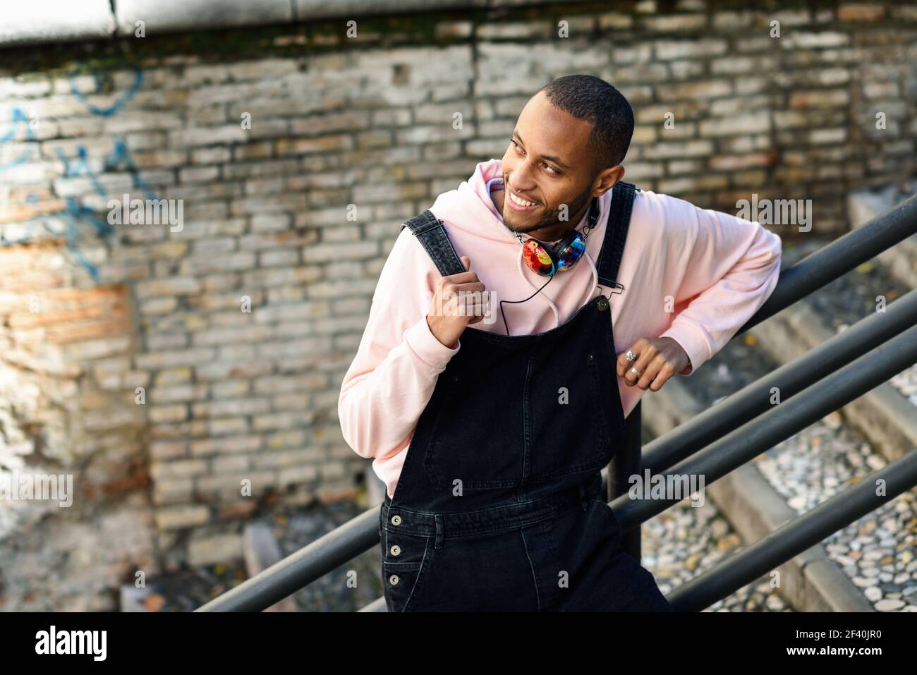 Giovane uomo nero che indossa abiti casual sorridendo sulle scale urbane. Ragazzo africano felice con pantaloni di bib all'aperto. Uomo nero sorridente che indossa abiti casual all'aperto Foto Stock
