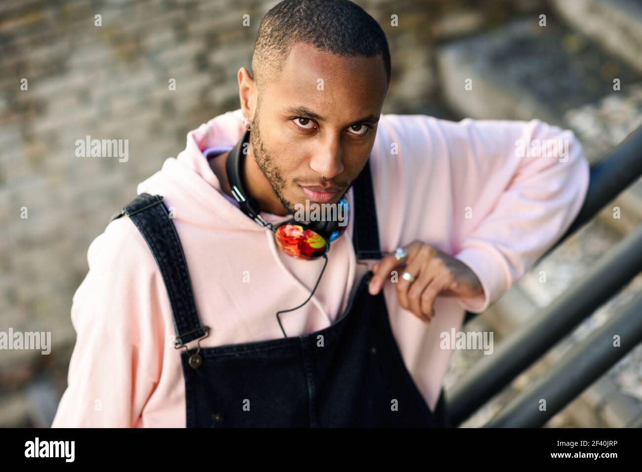 Giovane uomo nero che indossa abiti casual sui gradini urbani. Serio ragazzo africano con pantaloni di bib all'aperto. Serio ragazzo africano con pantaloni di bib all'aperto Foto Stock