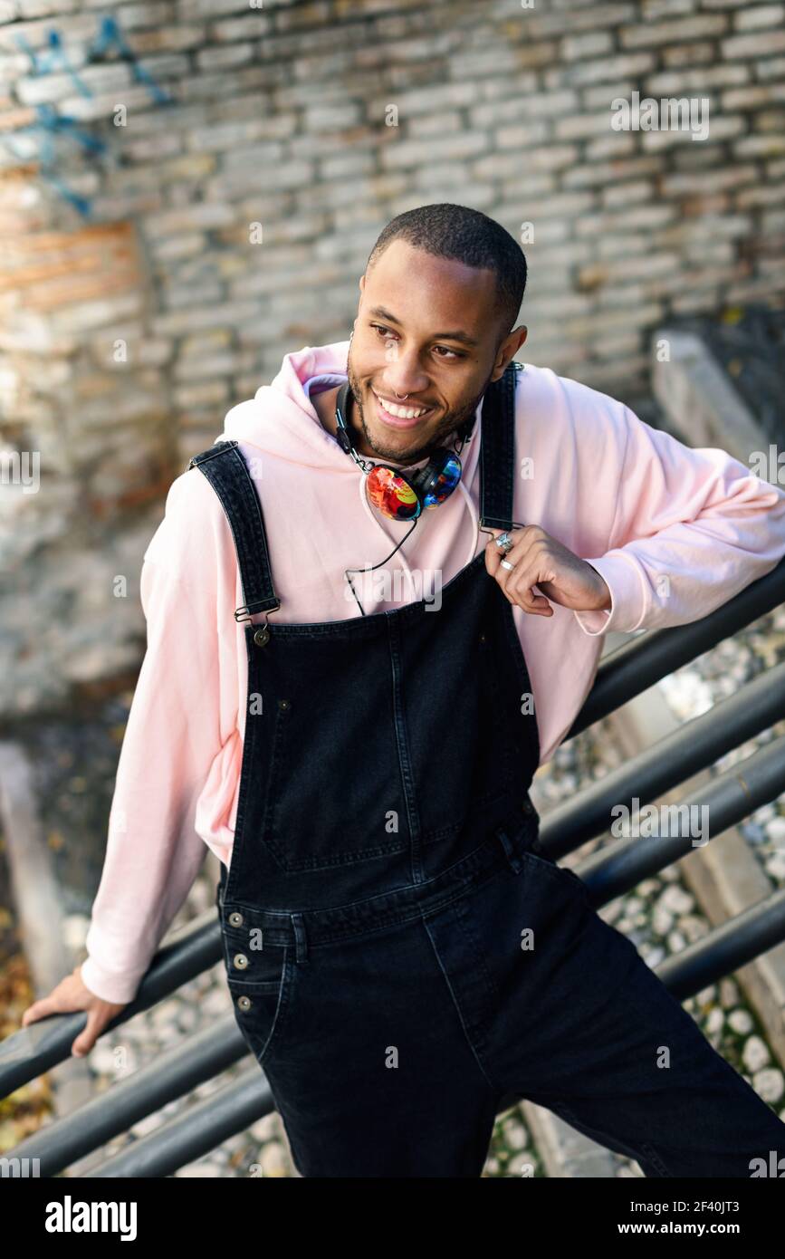 Giovane uomo nero che indossa abiti casual sorridendo sulle scale urbane. Ragazzo africano felice con pantaloni di bib all'aperto. Uomo nero sorridente che indossa abiti casual all'aperto Foto Stock