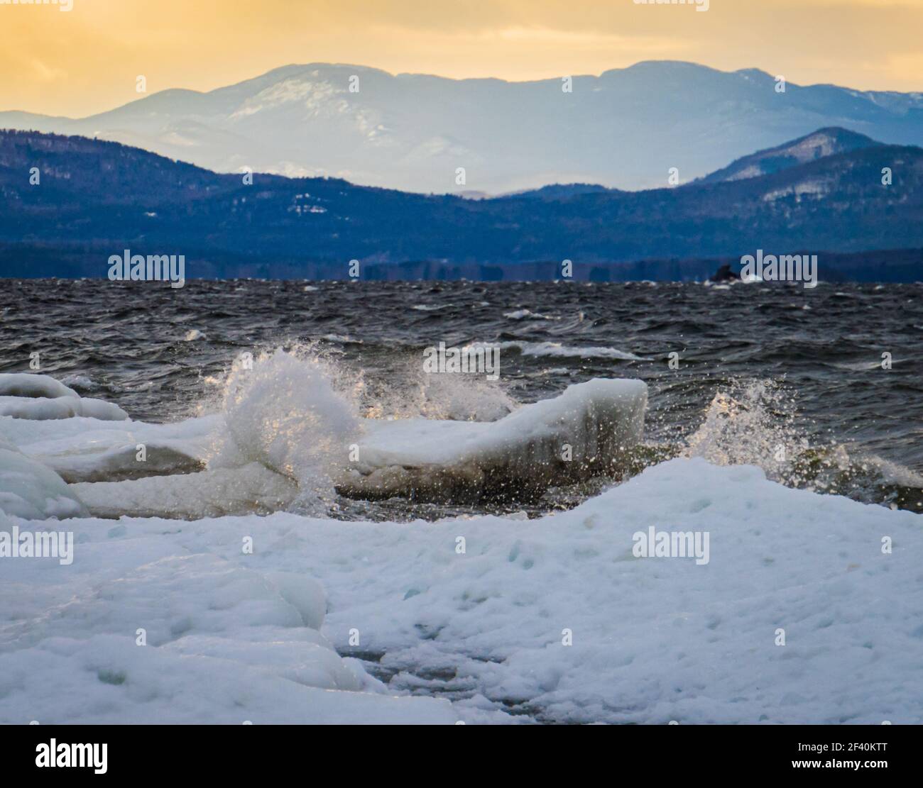 Onde di luce del tramonto che si infrangono sulla costa ghiacciata e fredda del lago Champlain con le montagne Adirondack a New York attraverso lago Foto Stock